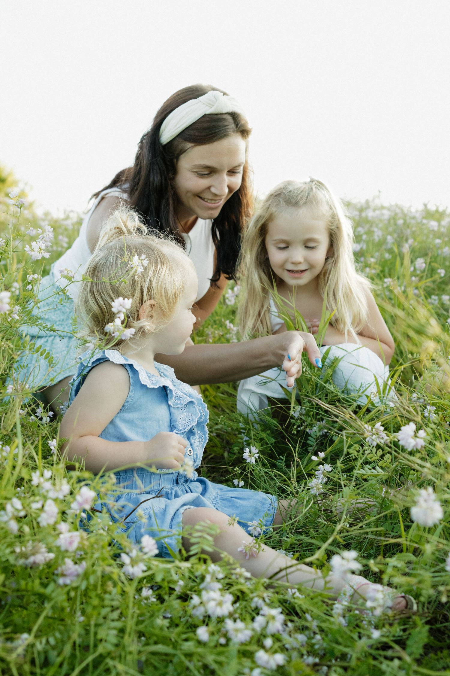 A smiling mother sits in a wildflower meadow with her two young blonde daughters, picking small white flowers together.