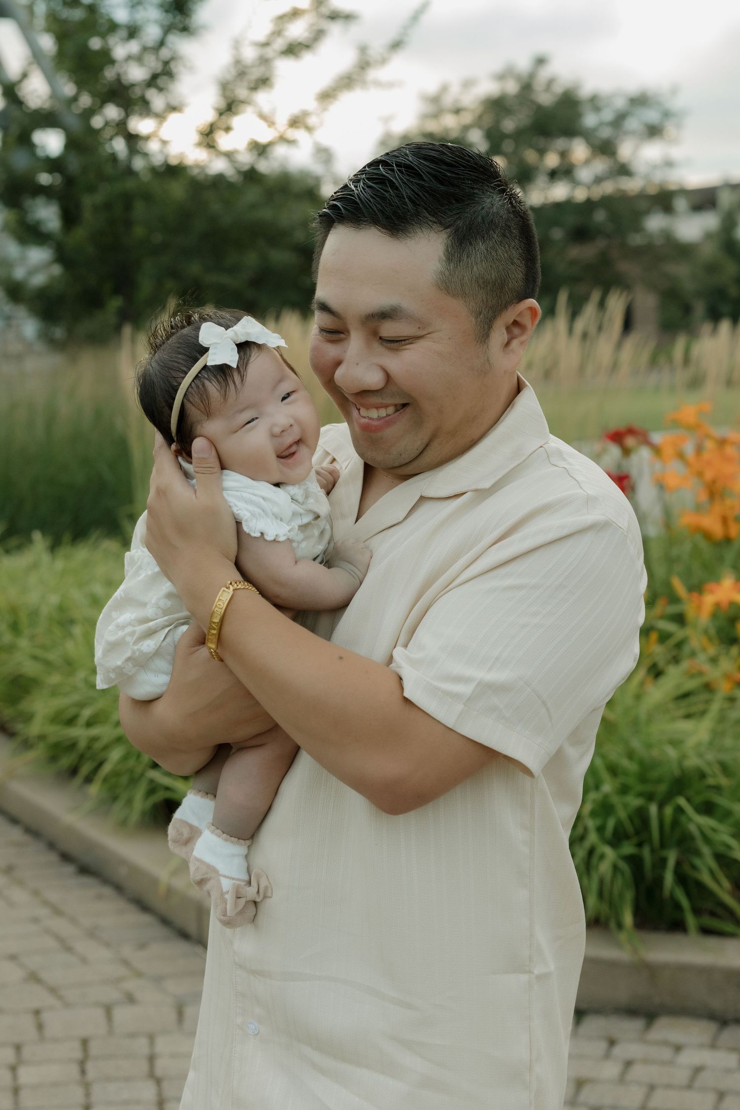 Smiling Asian man holding a happy baby girl in a white dress outdoors in a lush garden setting.
