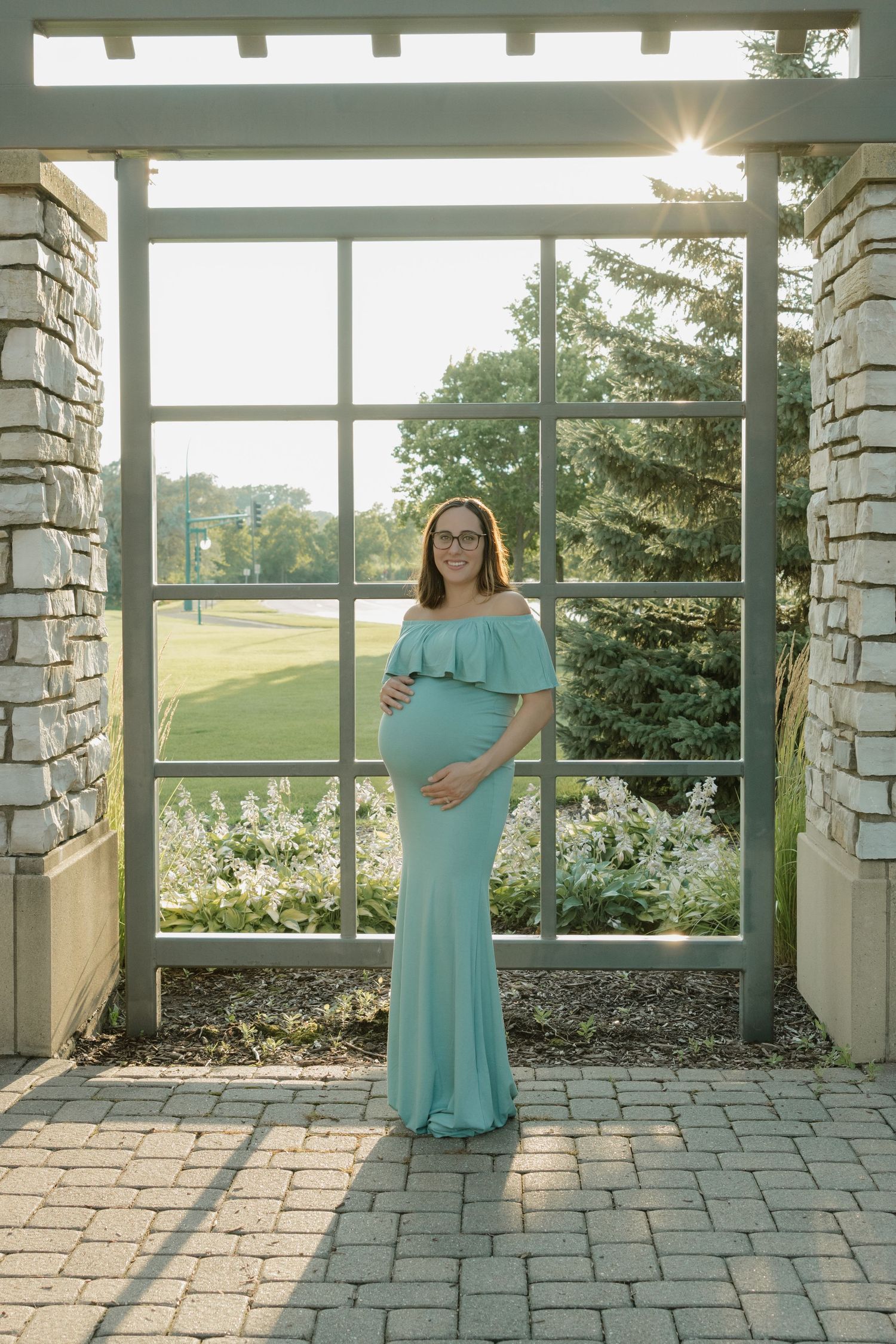 Pregnant woman in teal off-shoulder gown standing in garden pergola, cradling baby bump in golden sunlight.