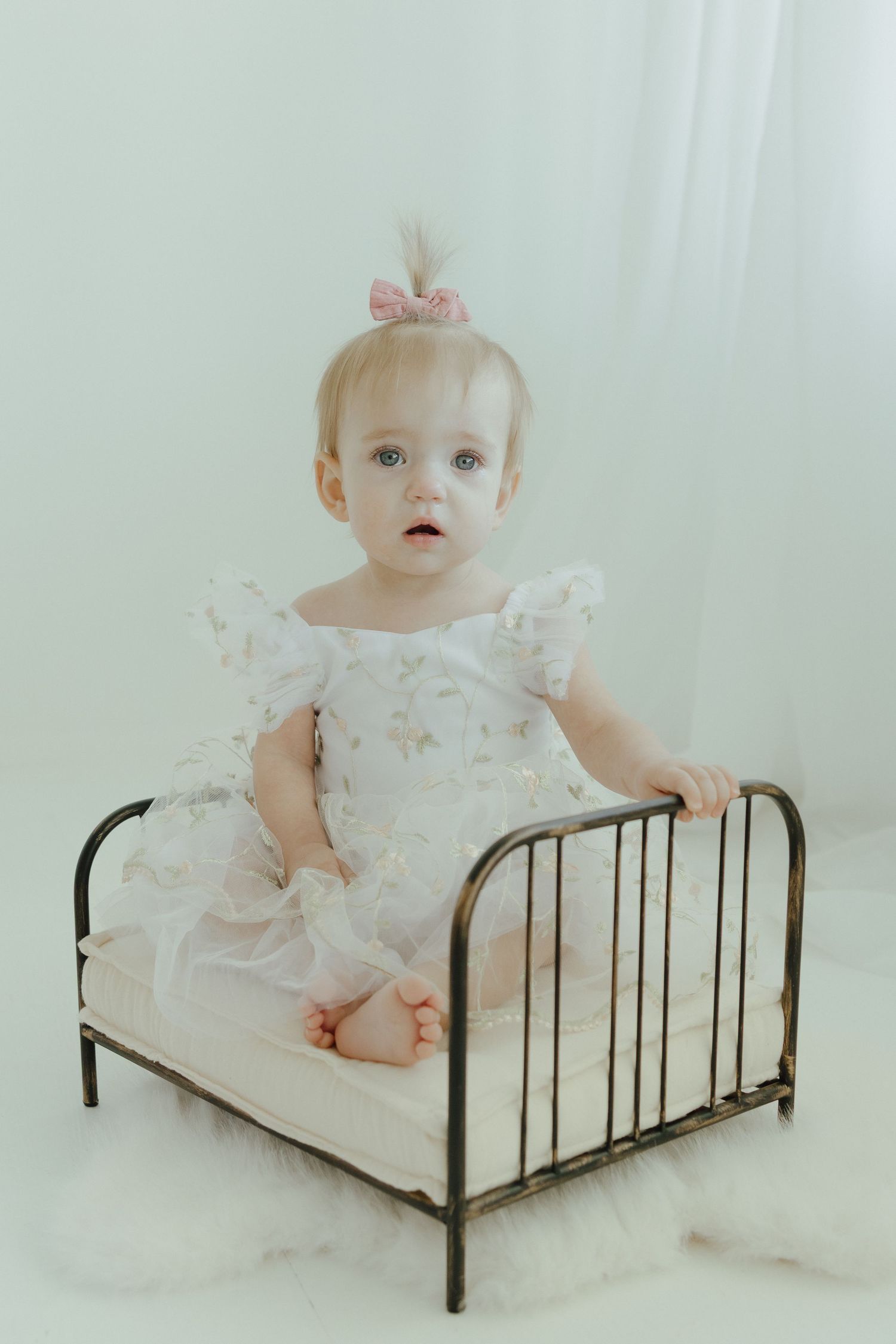 Baby in white lace dress crawls and plays on antique metal bed frame against light background.
