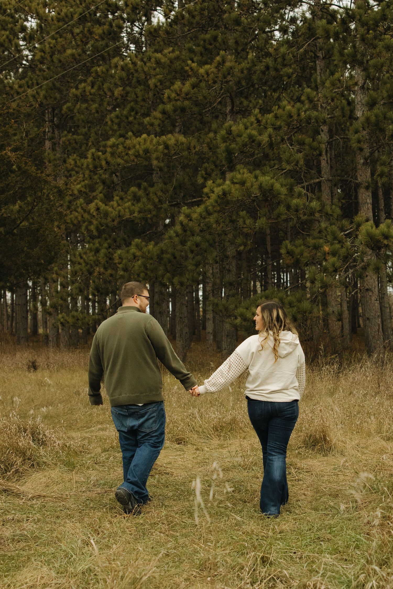 Couple holding hands walking through golden field toward tall pine forest in autumn, viewed from behind.