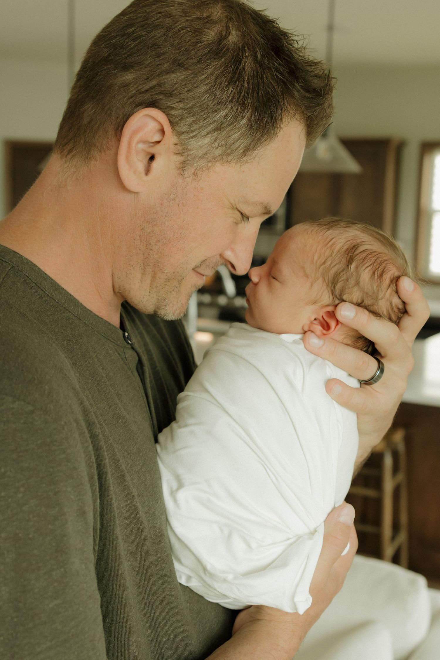 A father tenderly holds his swaddled newborn baby close, touching noses in an intimate indoor moment.