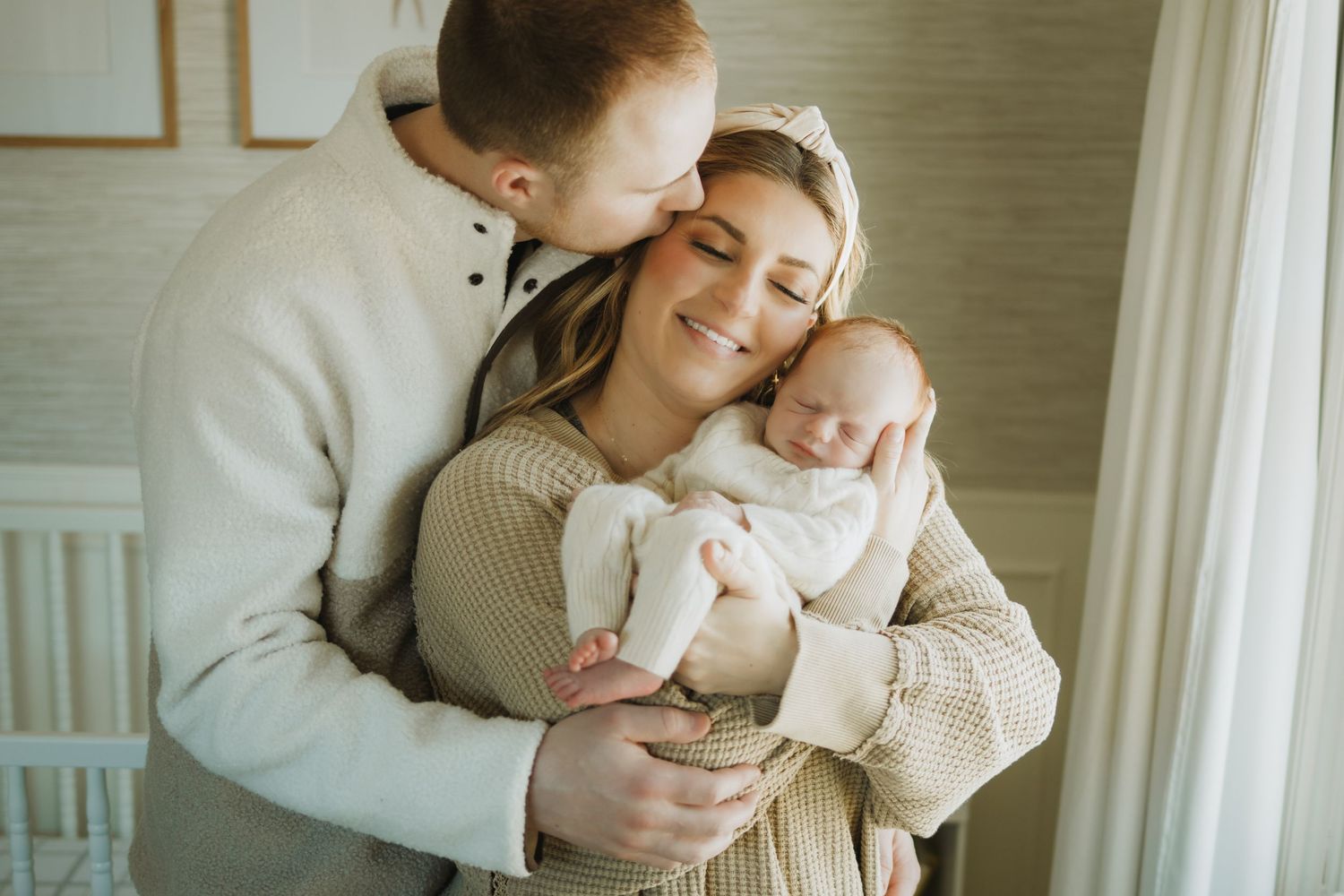 Happy couple smiling and kissing while lovingly cradling their newborn baby in a bright, cozy nursery room.