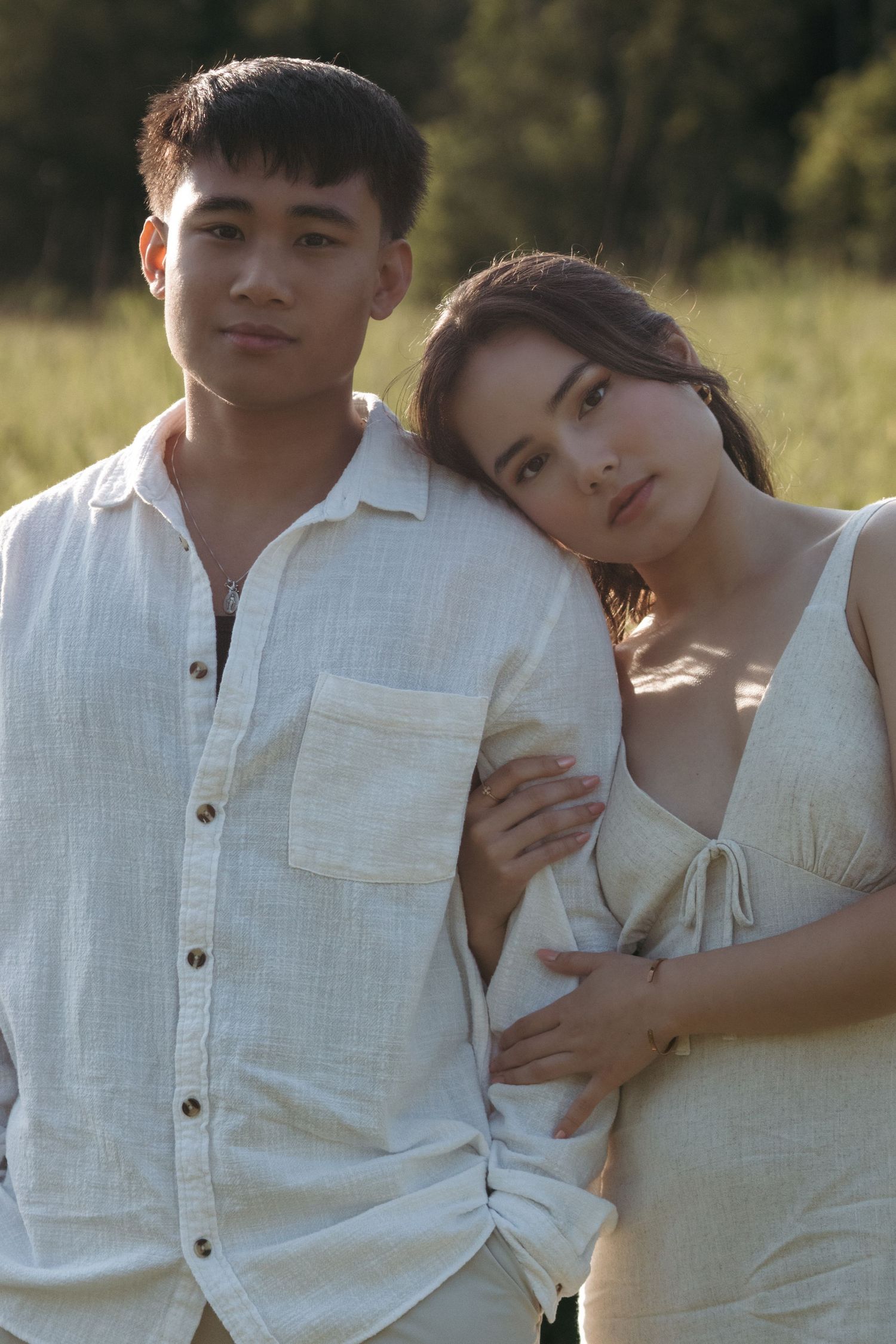 A romantic couple in white outfits poses together in a sunlit field during a golden hour photoshoot.