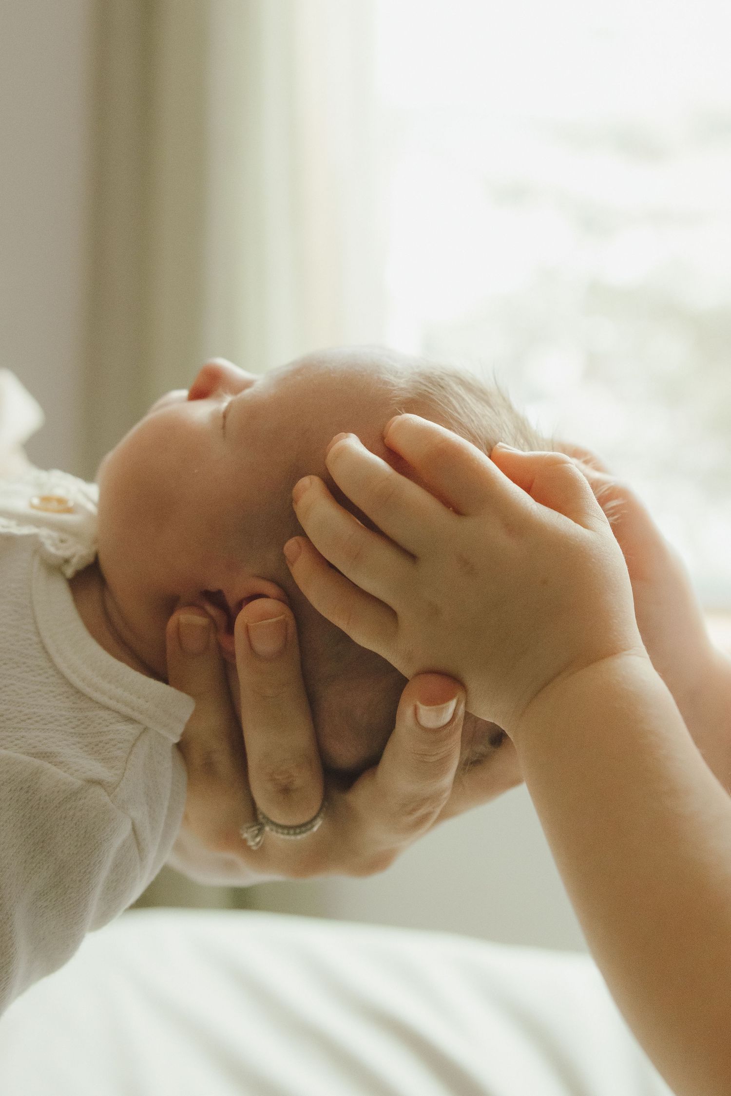 Adult hands gently cradling a sleeping newborn baby's head, with soft natural window light in the background.