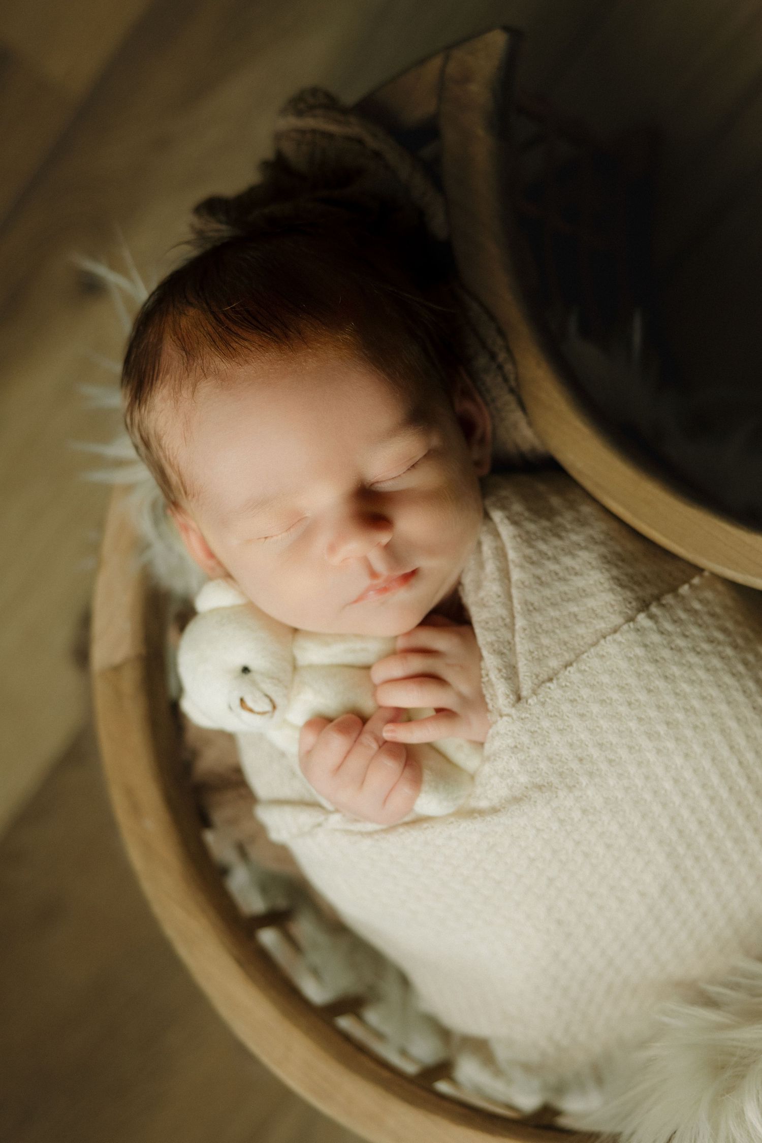 Newborn baby sleeping peacefully in a wooden bowl, wrapped in a white knit blanket, holding a small white stuffed animal.