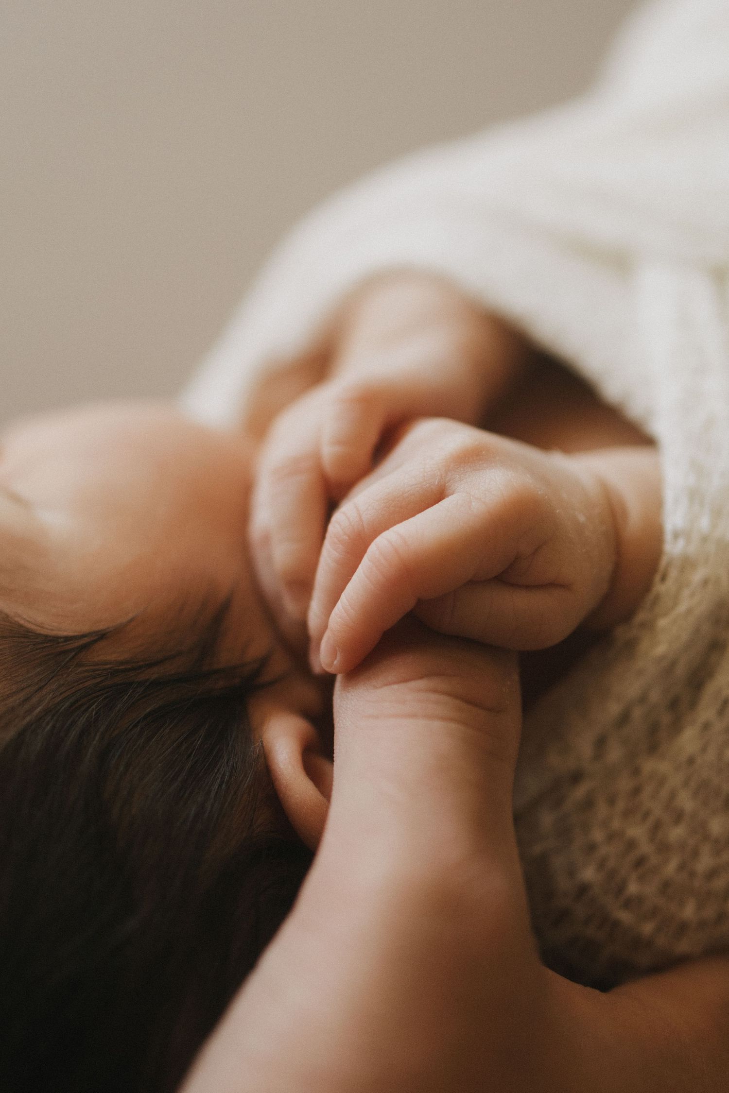 Close-up of a sleeping newborn baby gently holding an adult's finger, wrapped in a soft cream blanket.