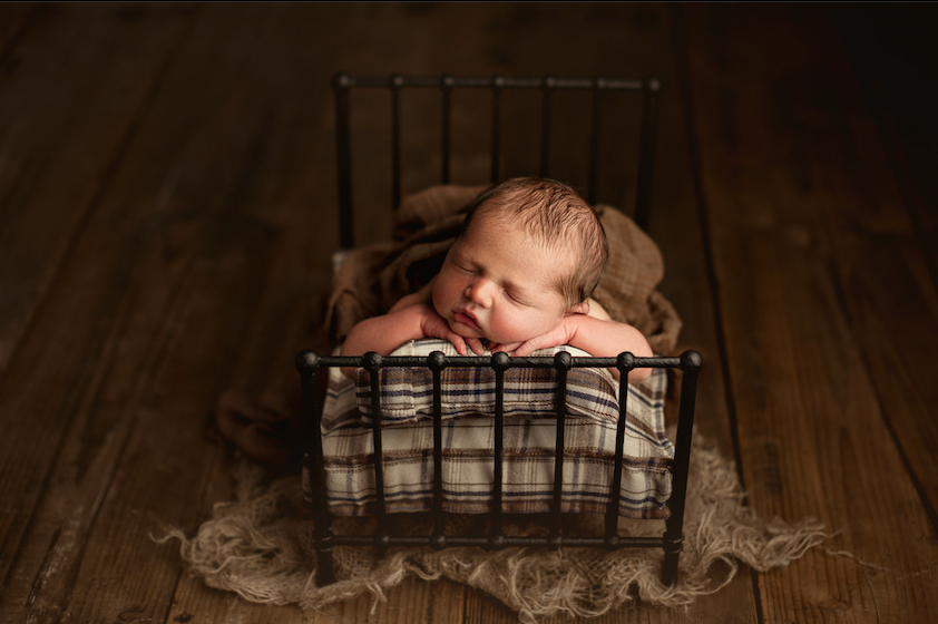 Newborn baby sleeping peacefully in a tiny vintage metal bed with plaid mattress on a rustic wooden floor.