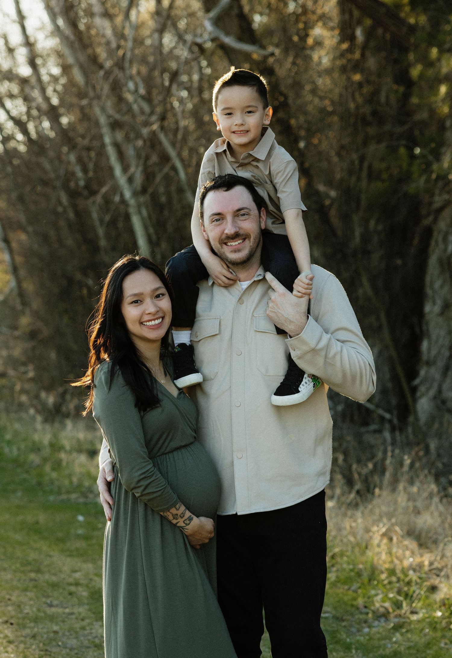Family portrait in nature with expecting mother in green dress and small child sitting on father's shoulders.