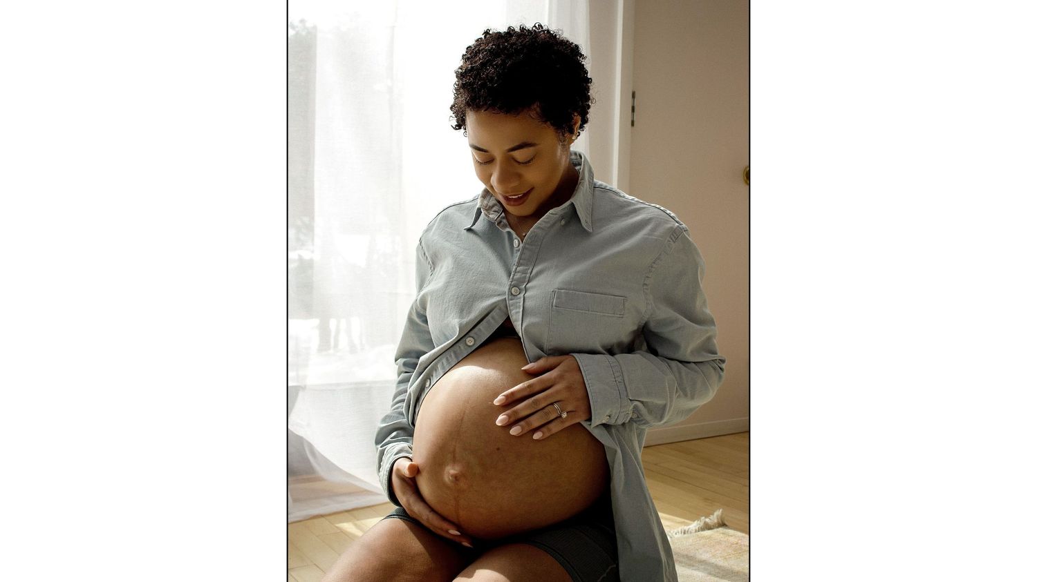 Candid maternity photo of person in denim shirt by window light.