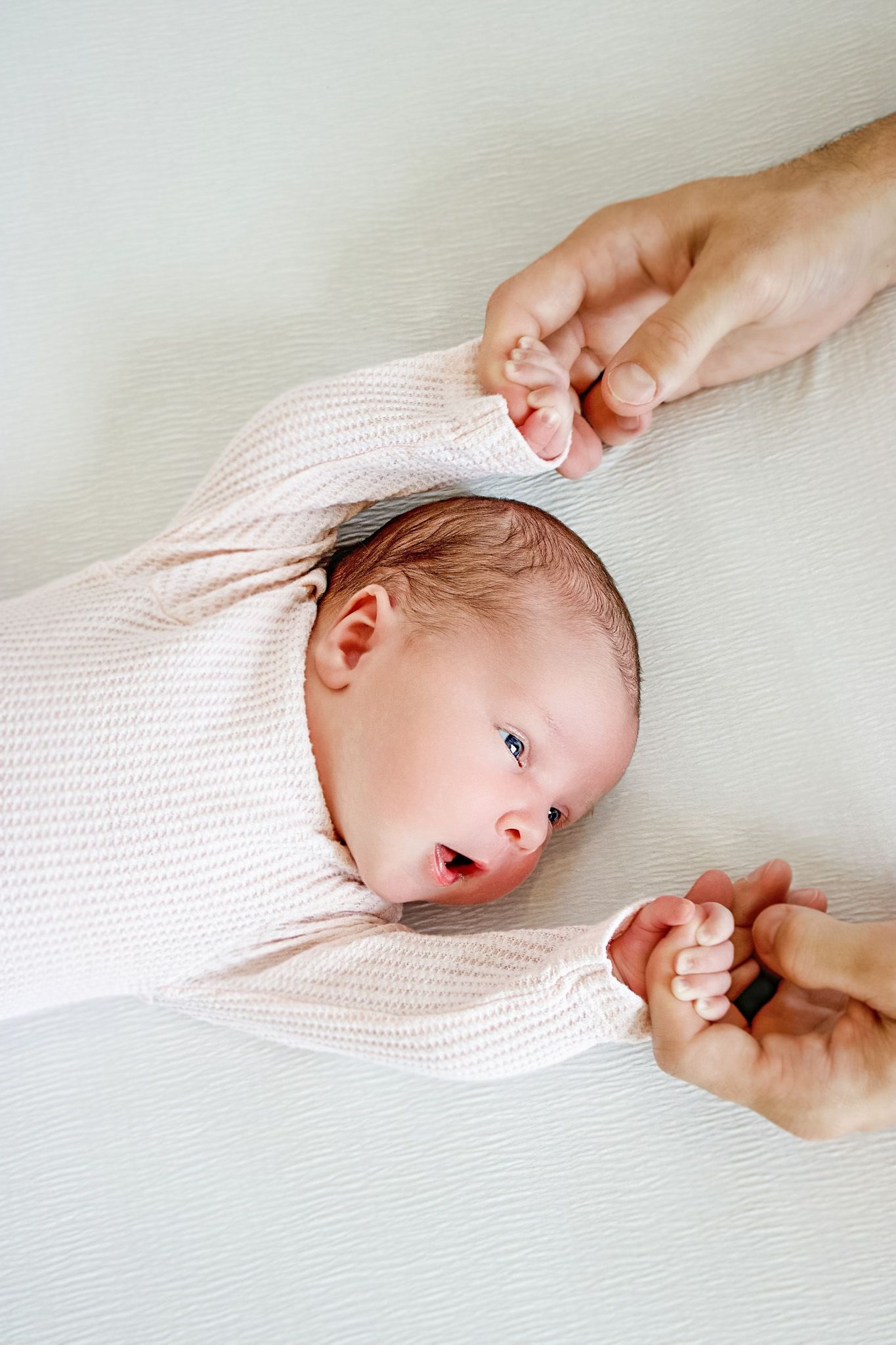 Newborn baby in pink onesie lying on white surface, held by adult hands on both sides.