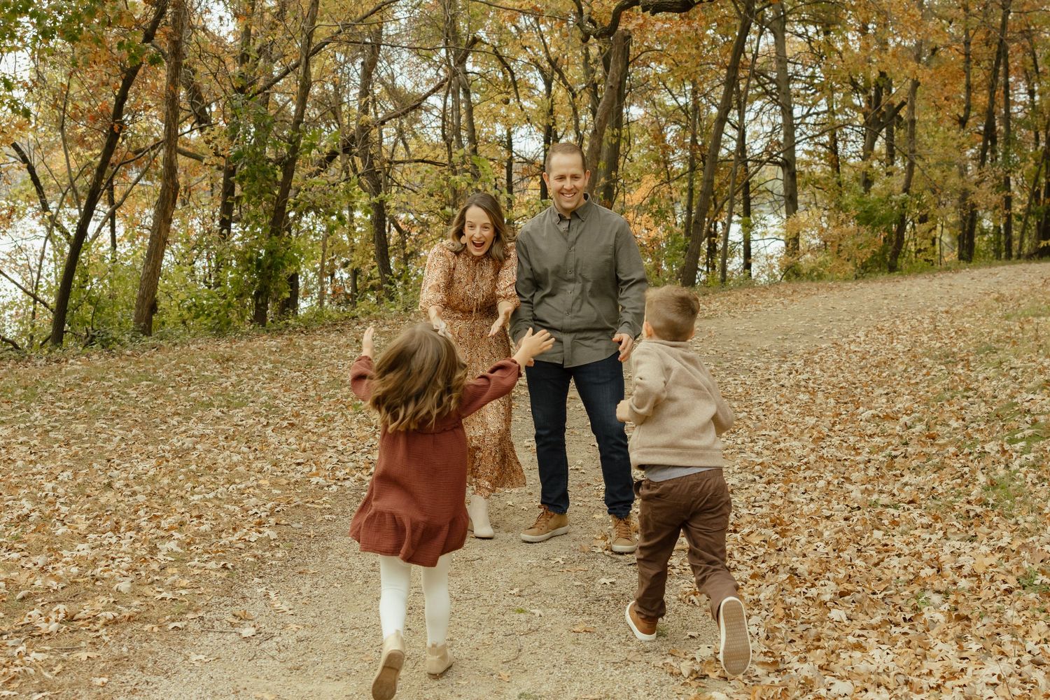 Family of four playing outdoors in autumn forest with fallen leaves, parents watching children run on a scenic trail.