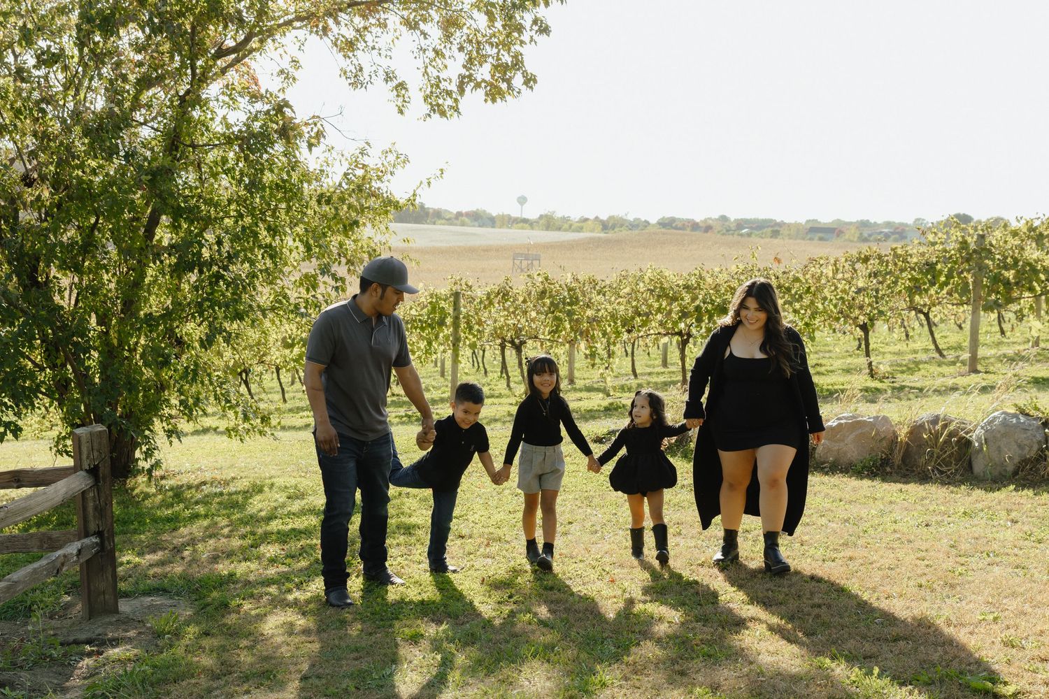 Family of five holding hands walking through a lush vineyard on a sunny day.