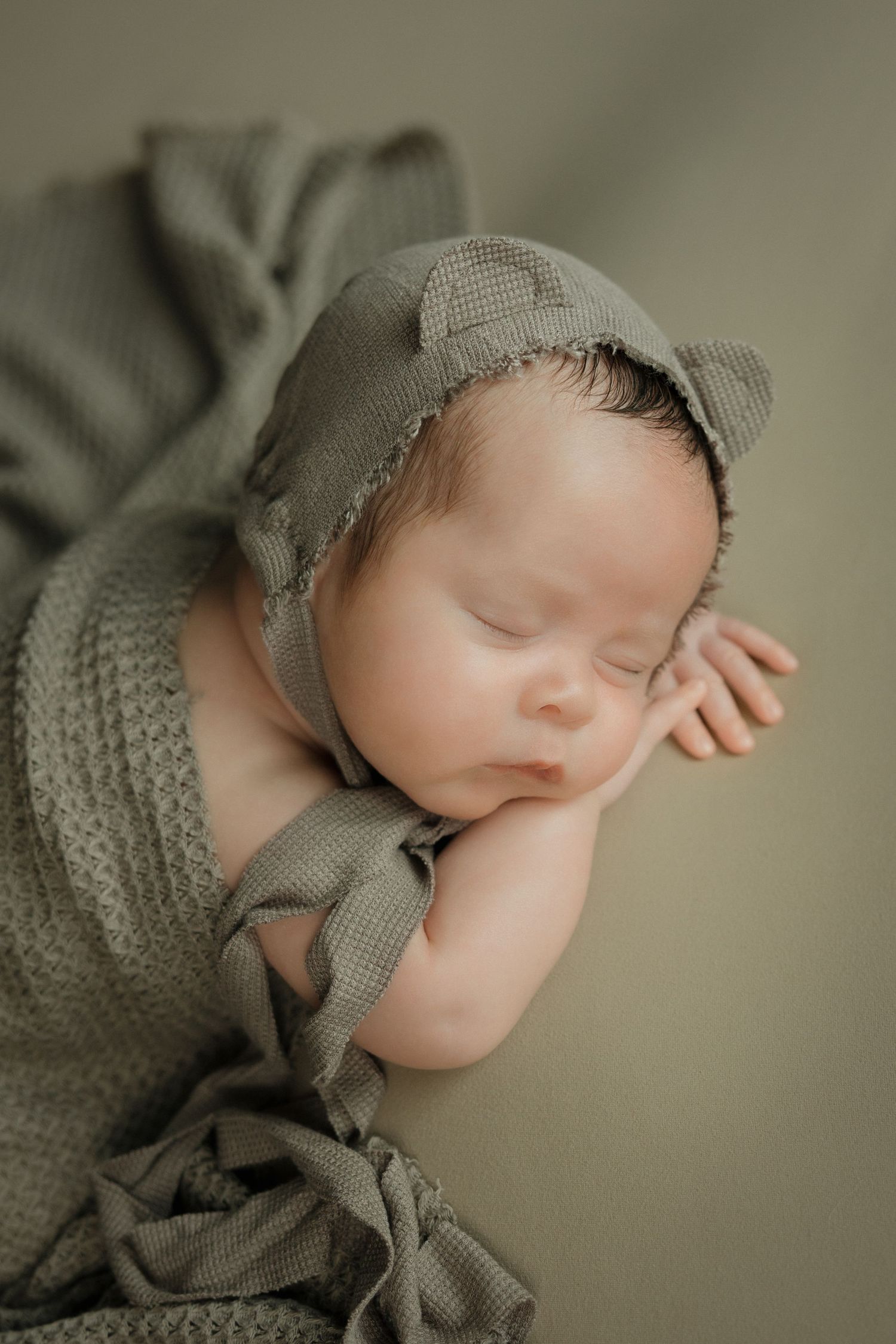 Peaceful newborn baby sleeping in sage green bonnet and wrap on matching green background.