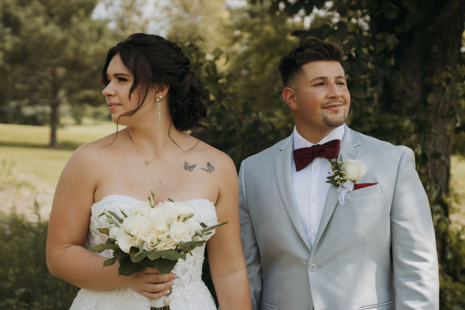 Bride holding white bouquet stands beside groom in silver suit with burgundy bow tie outdoors at wedding ceremony.