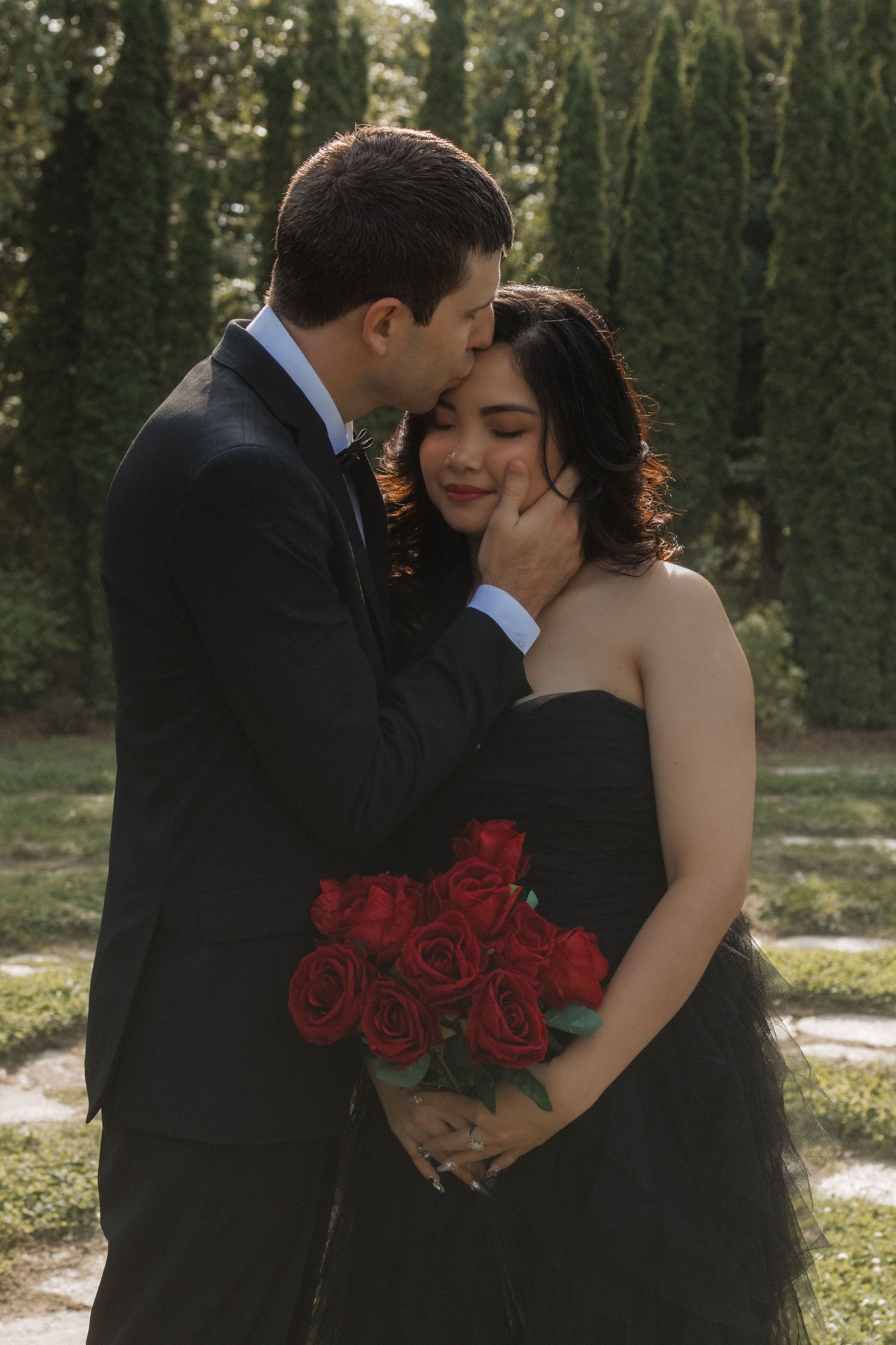 A romantic couple in formal wear embraces while holding red roses in a garden setting.