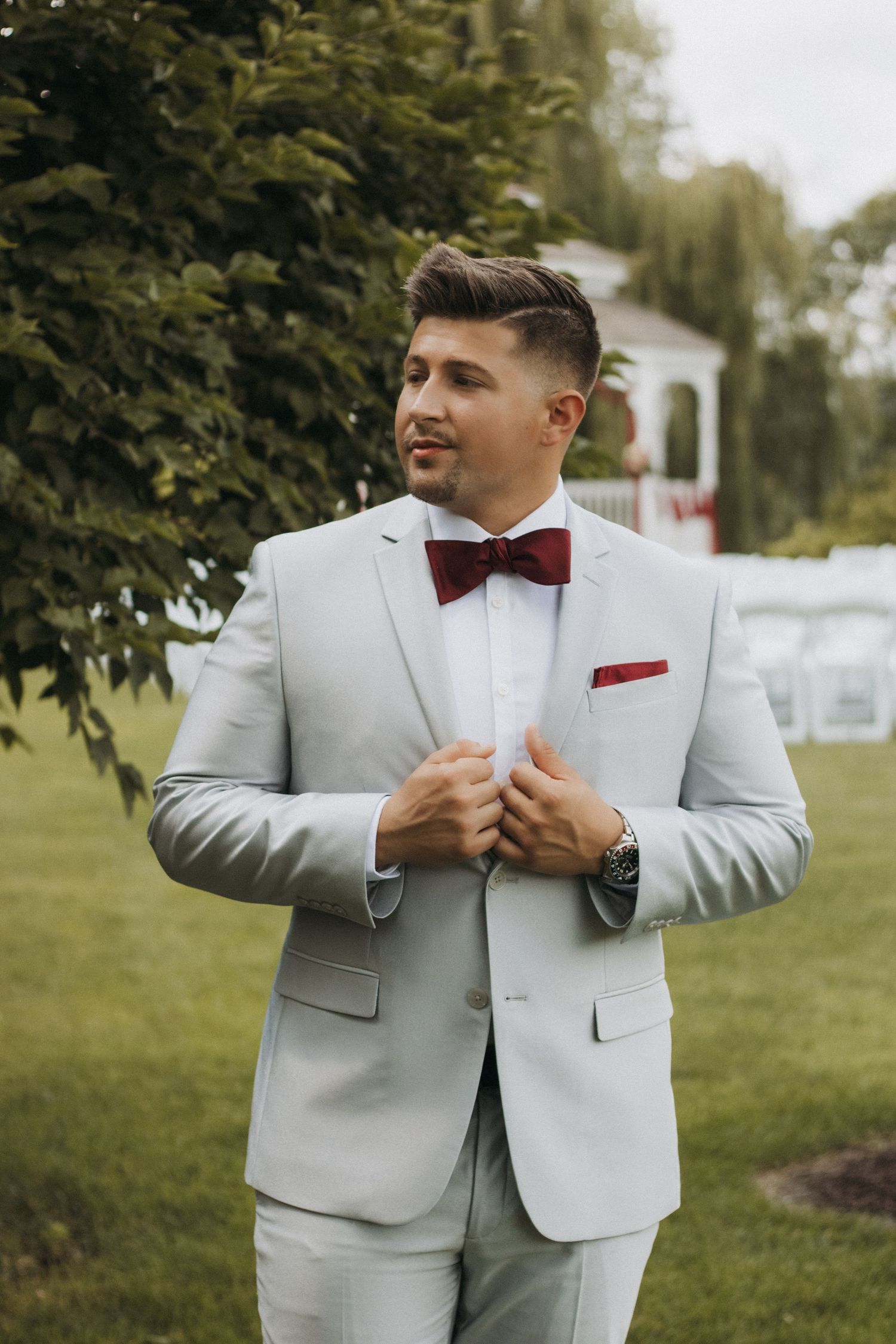 A person in a light gray suit and burgundy bow tie adjusts their attire while standing outdoors in a garden setting.