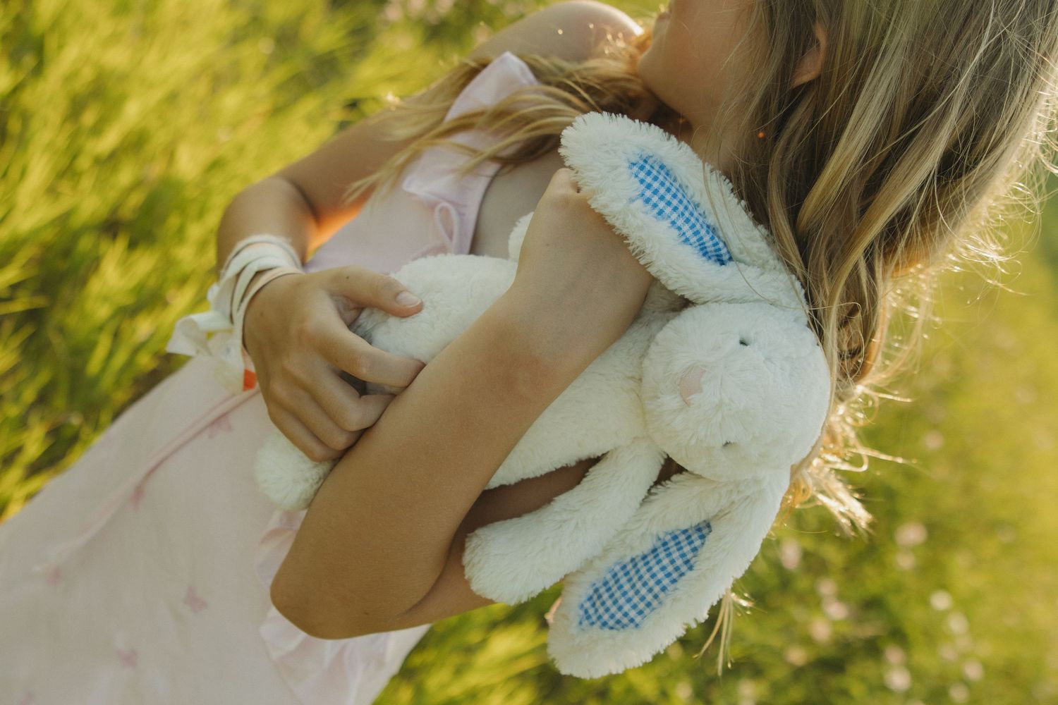 Young girl in pink dress hugging a white stuffed bunny rabbit toy while standing in a sunny green field.