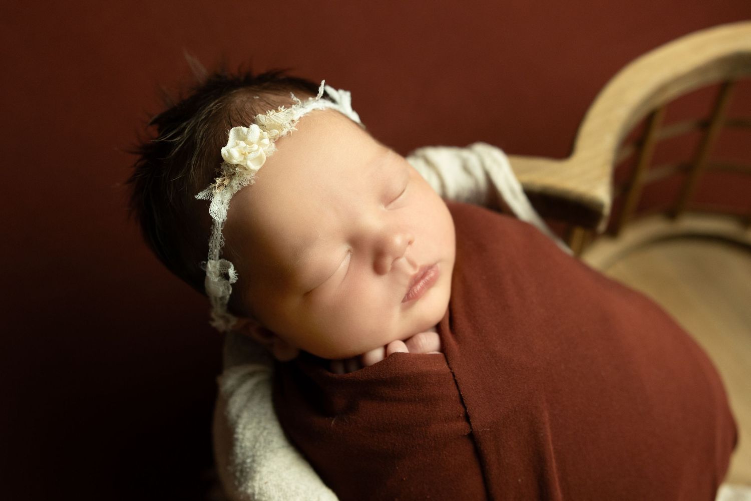 Sleeping newborn baby girl swaddled in burgundy wrap with white floral headband in a cozy bowl prop.
