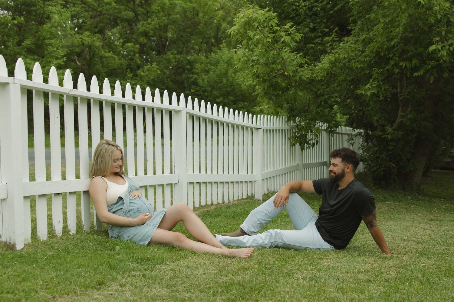 A couple sitting on green grass in a backyard, relaxing by a white picket fence surrounded by lush trees.