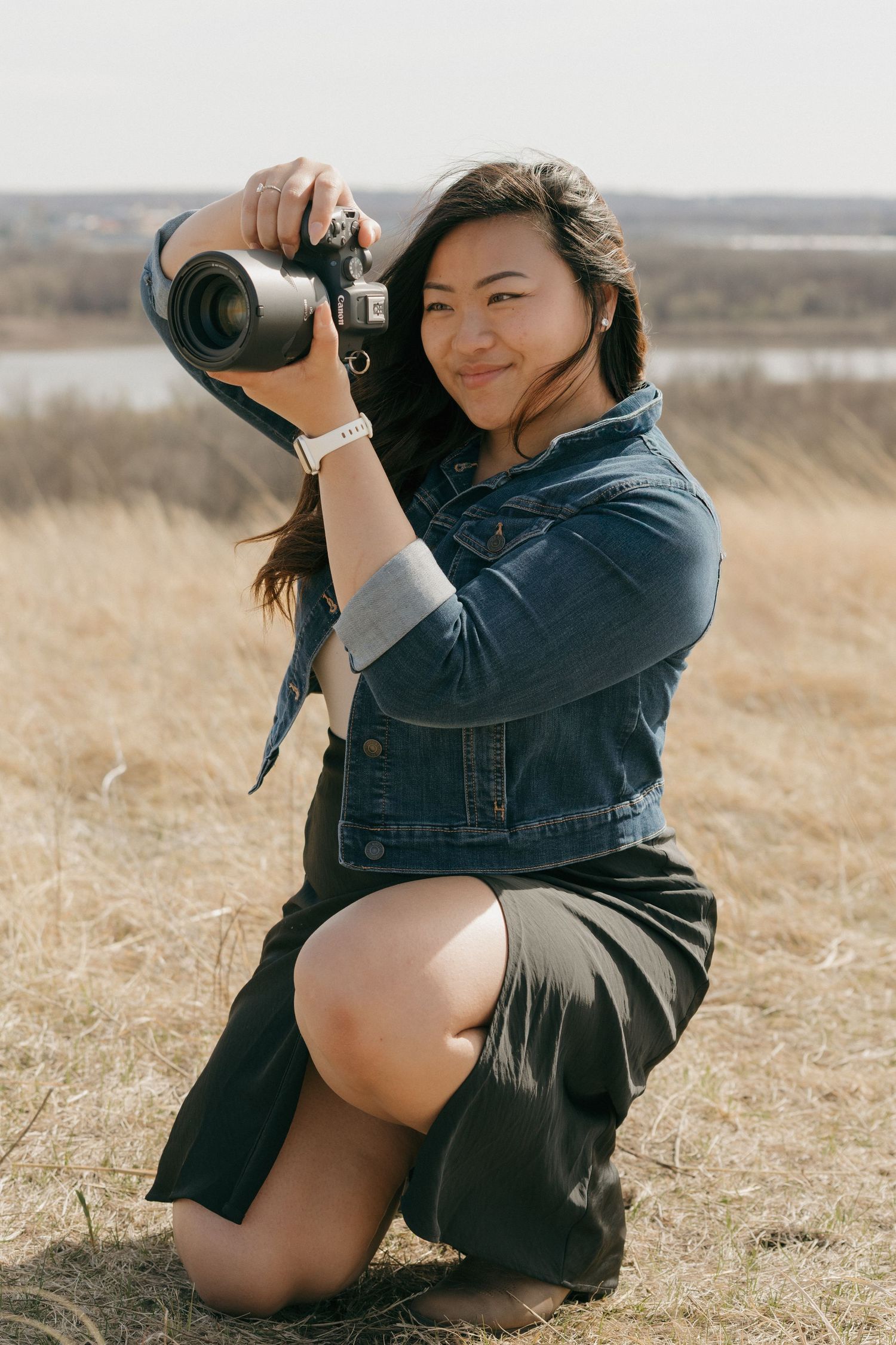 Photographer kneeling in dry grass field wearing denim jacket and skirt while using professional camera on sunny day.