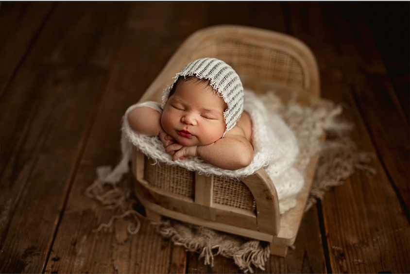 Sleeping newborn baby in knit bonnet nestled in rustic wooden bed with white lace wrap on dark wood floor.