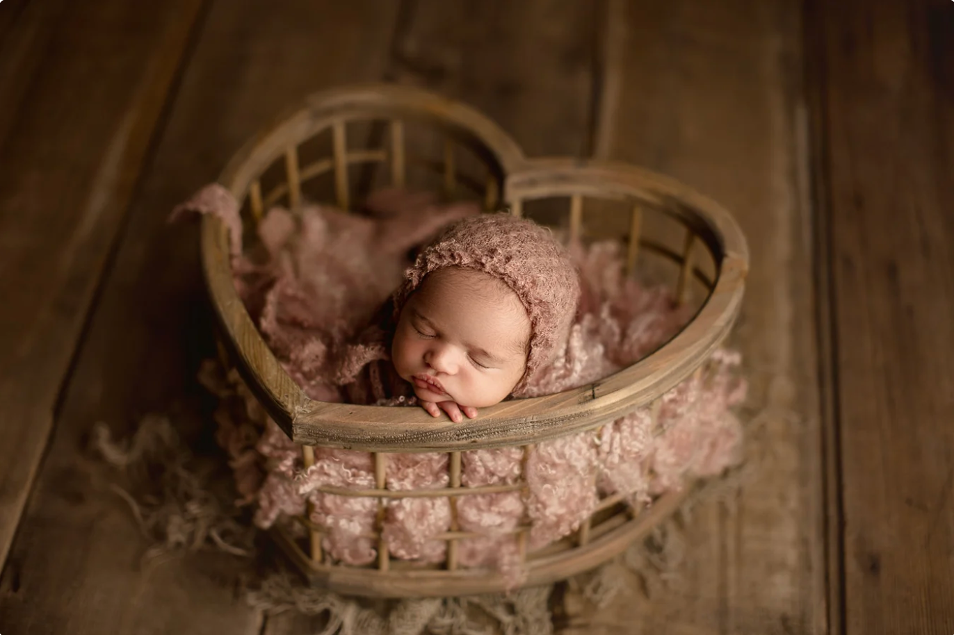 Newborn baby sleeping peacefully in a heart-shaped wicker basket with pink lace fabric and a knit bonnet.