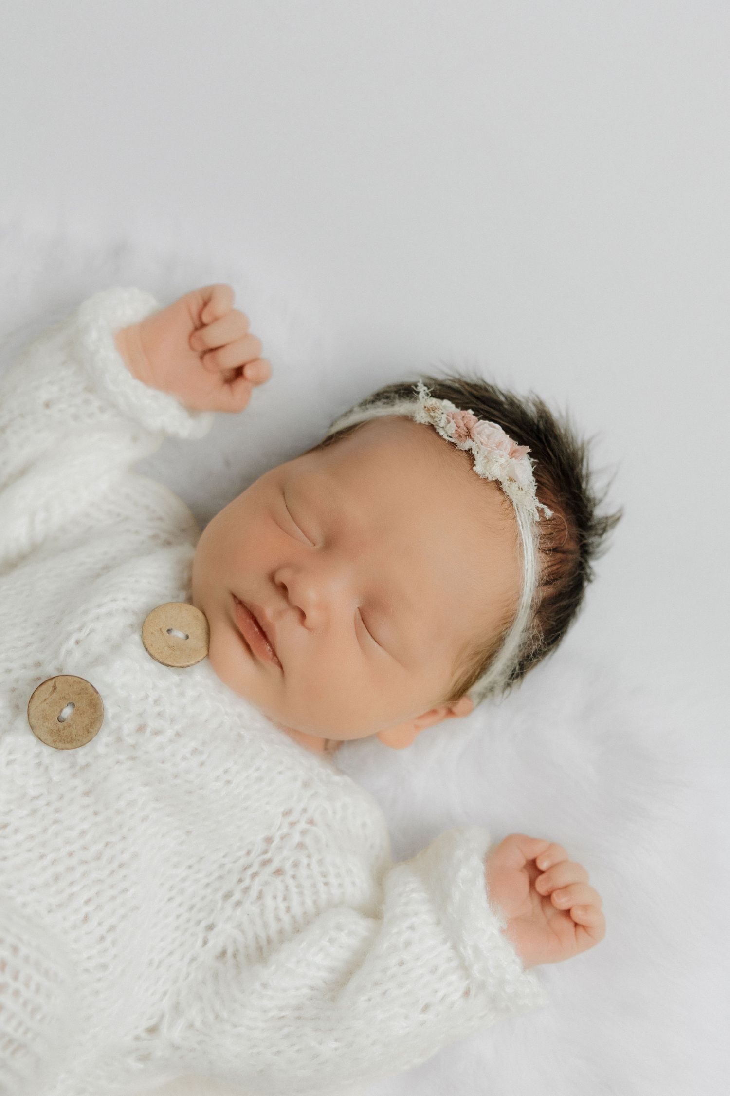 Sleeping newborn baby in white knit outfit with floral headband lying on soft white fur blanket.