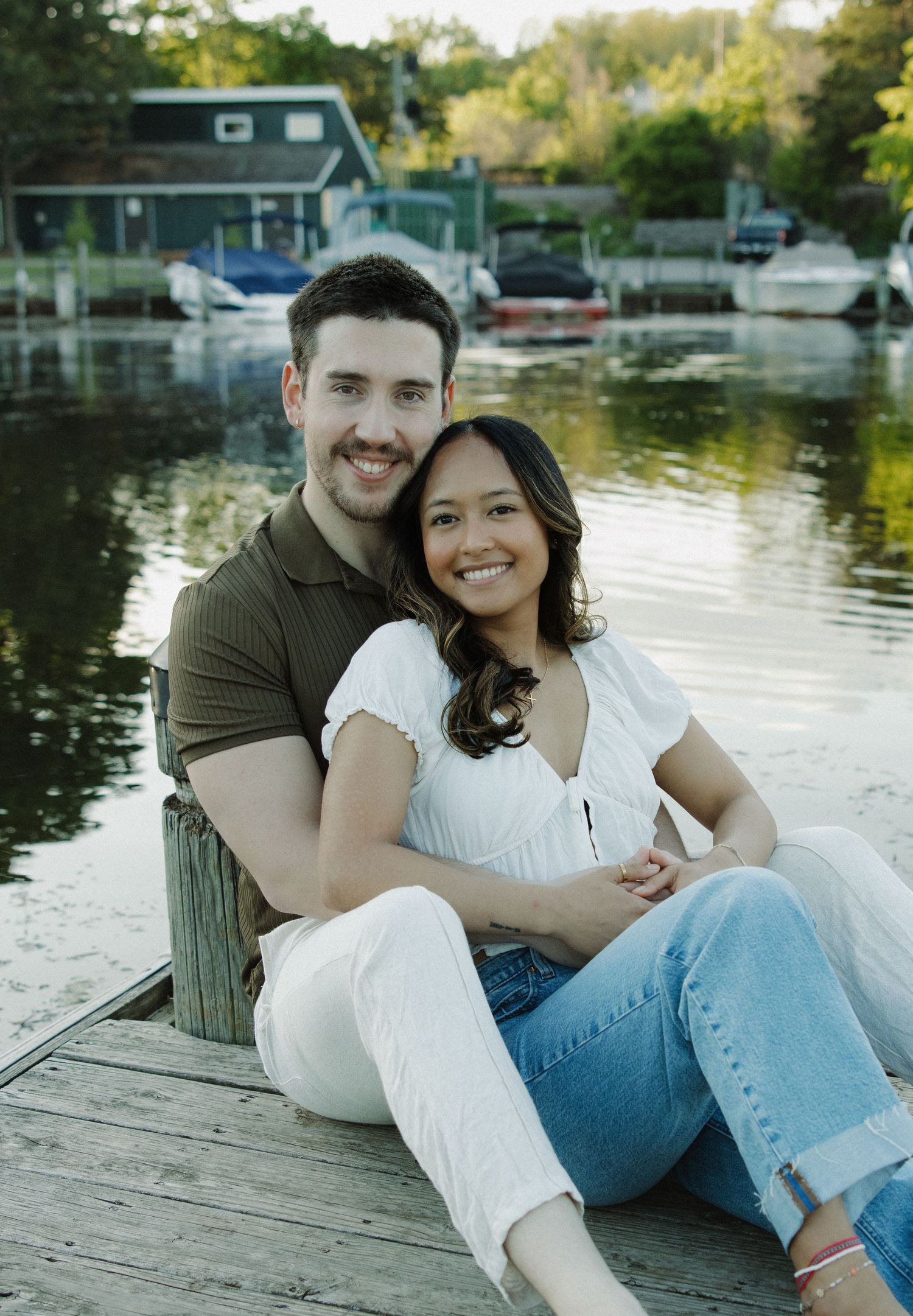 Couple sitting on wooden dock with boats and marina in background.