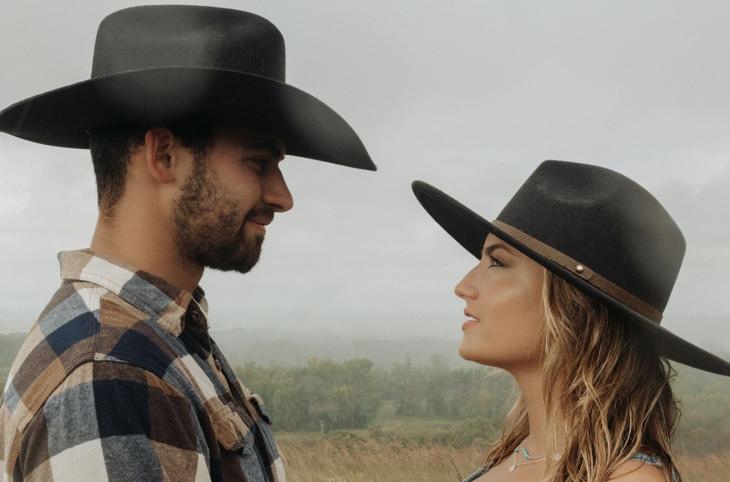 A couple in cowboy hats share an intimate moment in a rustic outdoor setting with soft lighting.