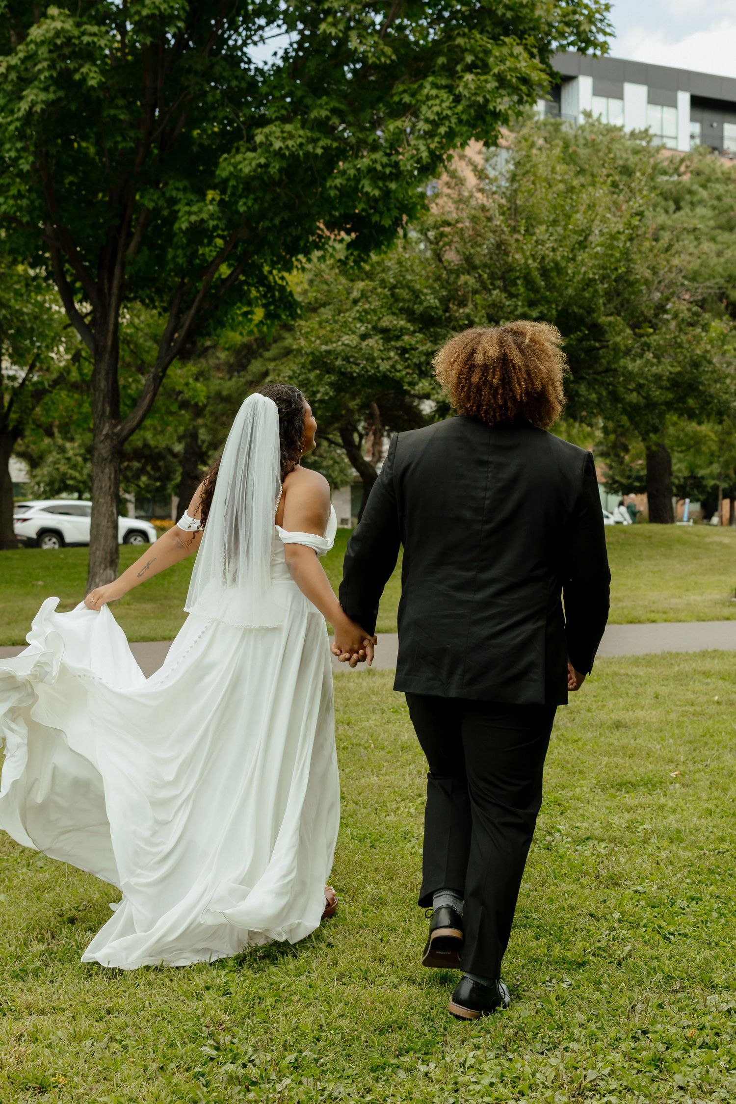 Bride in white gown and veil holds hands with groom in black suit walking through green park.
