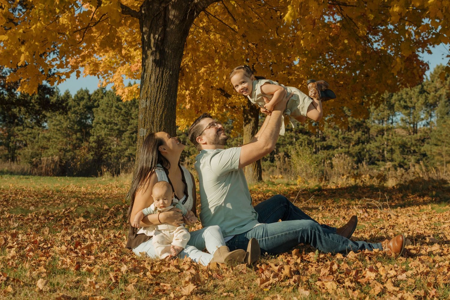 Family enjoying autumn park, parents playing with babies under golden maple tree on fallen leaves.