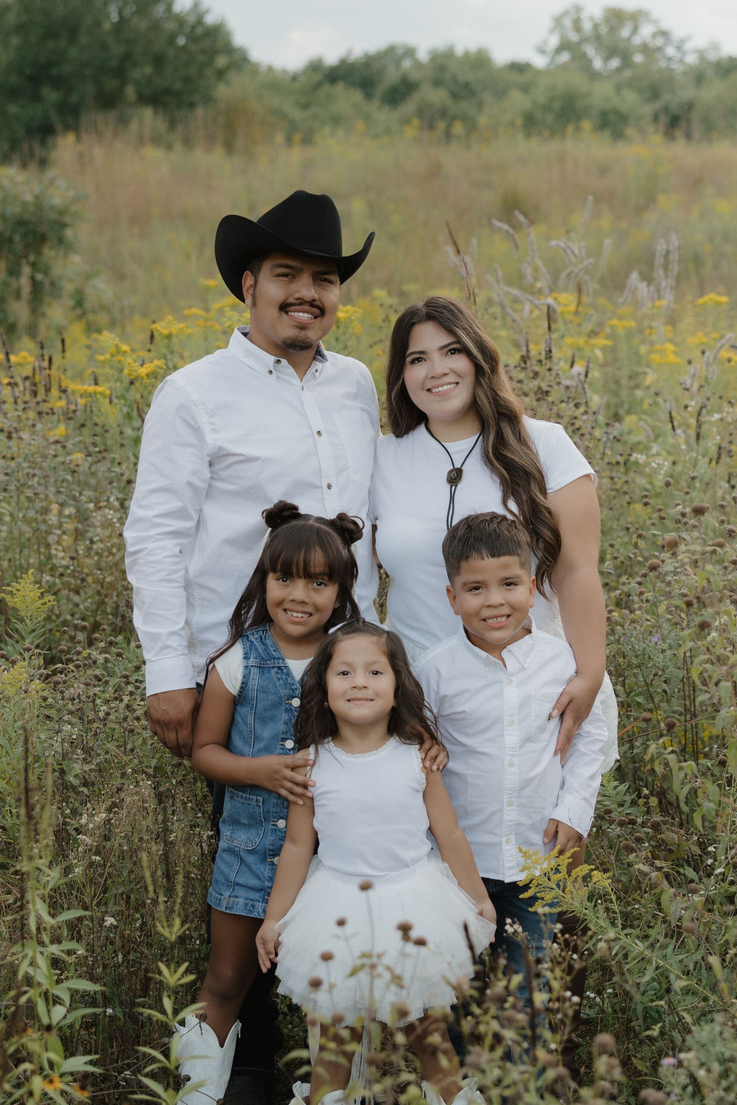 Happy family of five posing in a wildflower meadow, parents in white shirts with three young children.