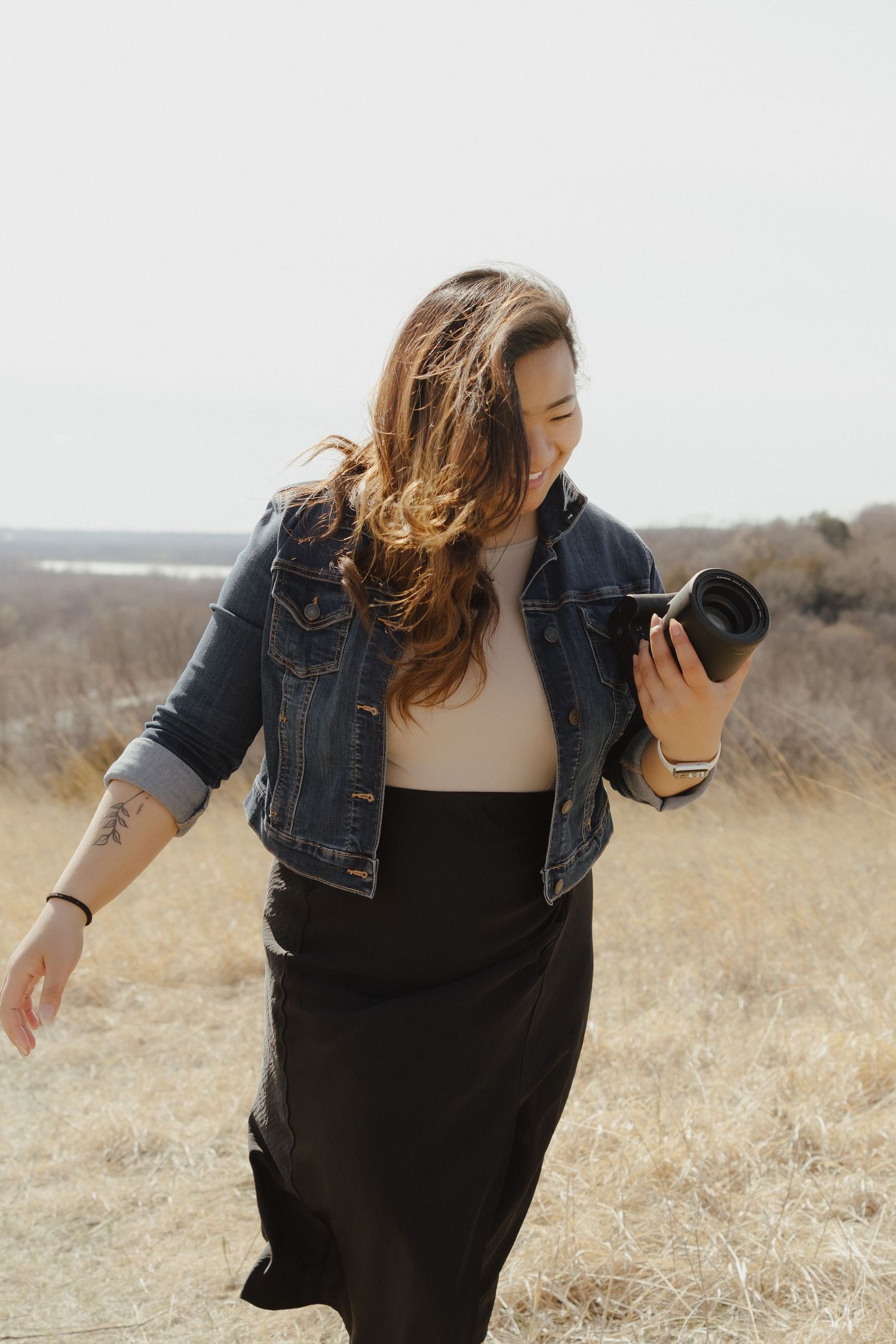 A woman in a black dress and denim jacket dances freely in a golden field during sunset.