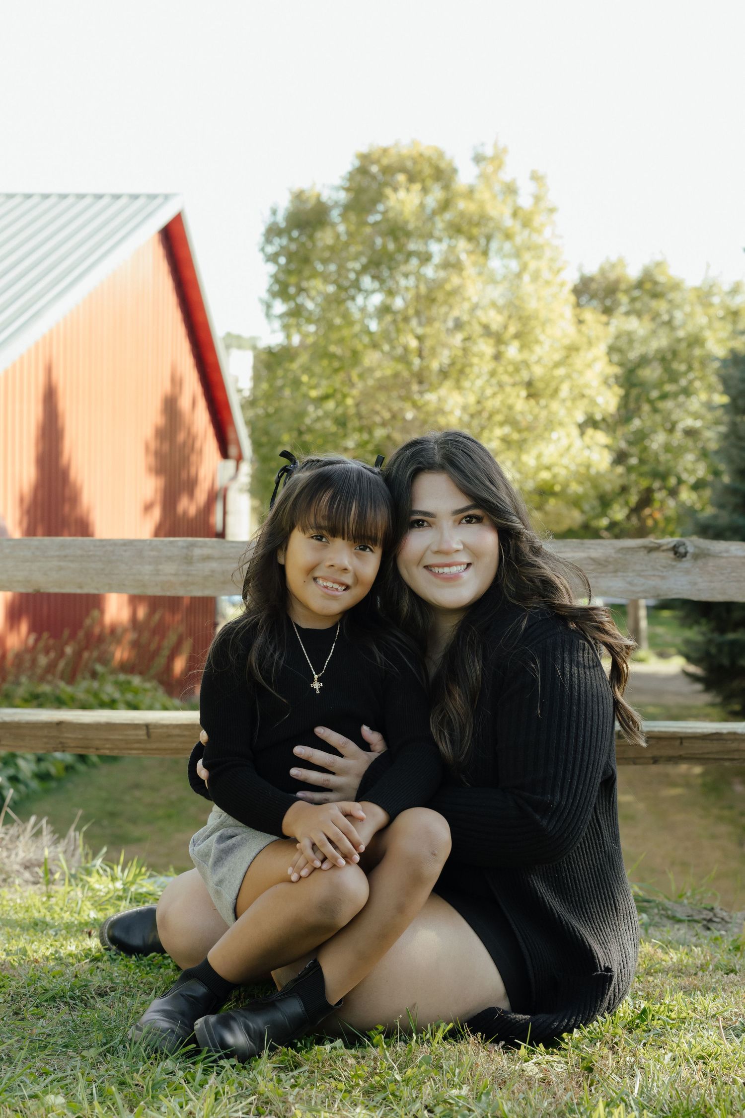 Two smiling girls sitting on grass in front of a wooden fence and red barn on a sunny day.