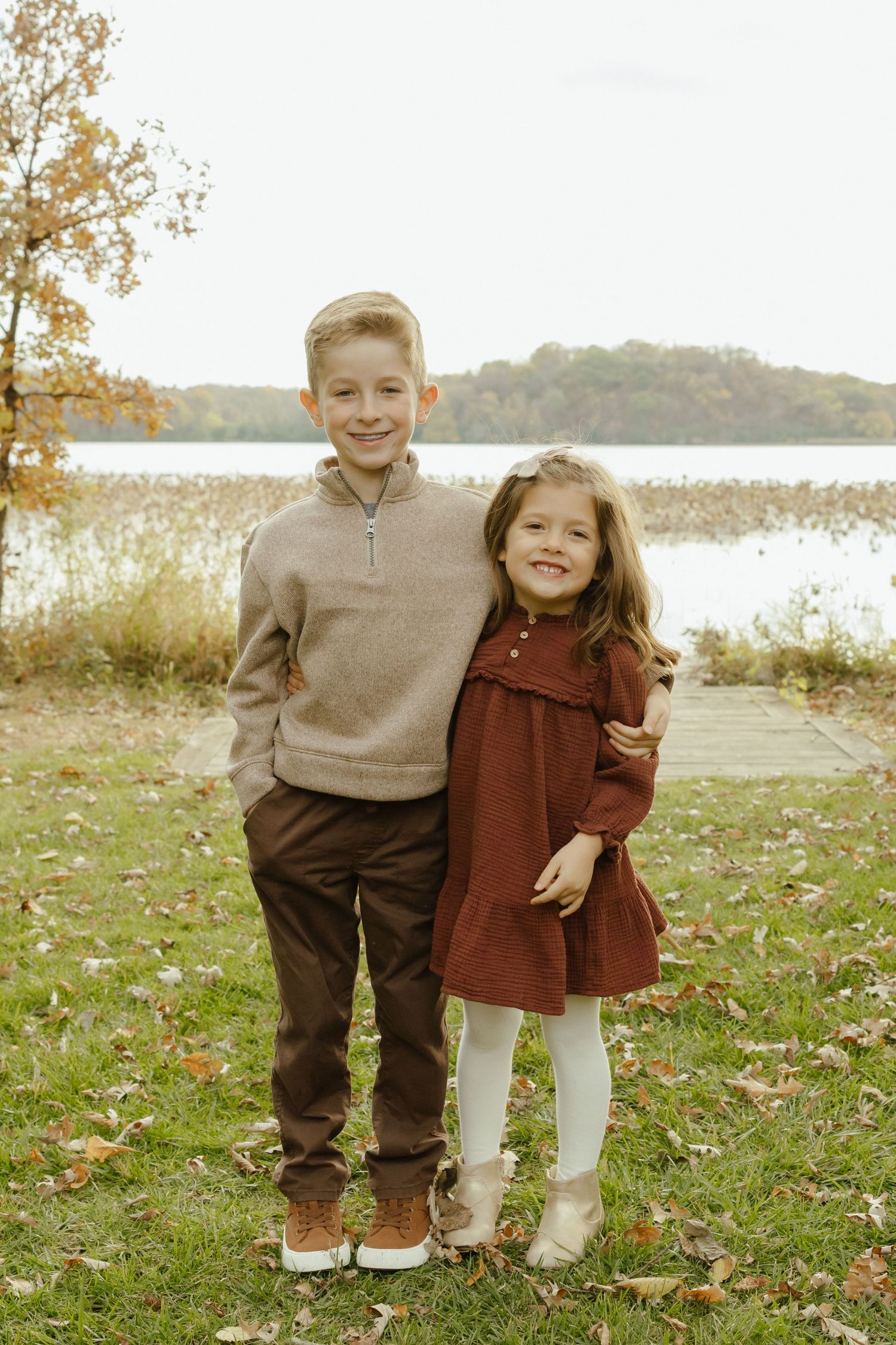 Two children pose outdoors near a lake in autumn, boy in tan sweater and girl in red dress smiling together.