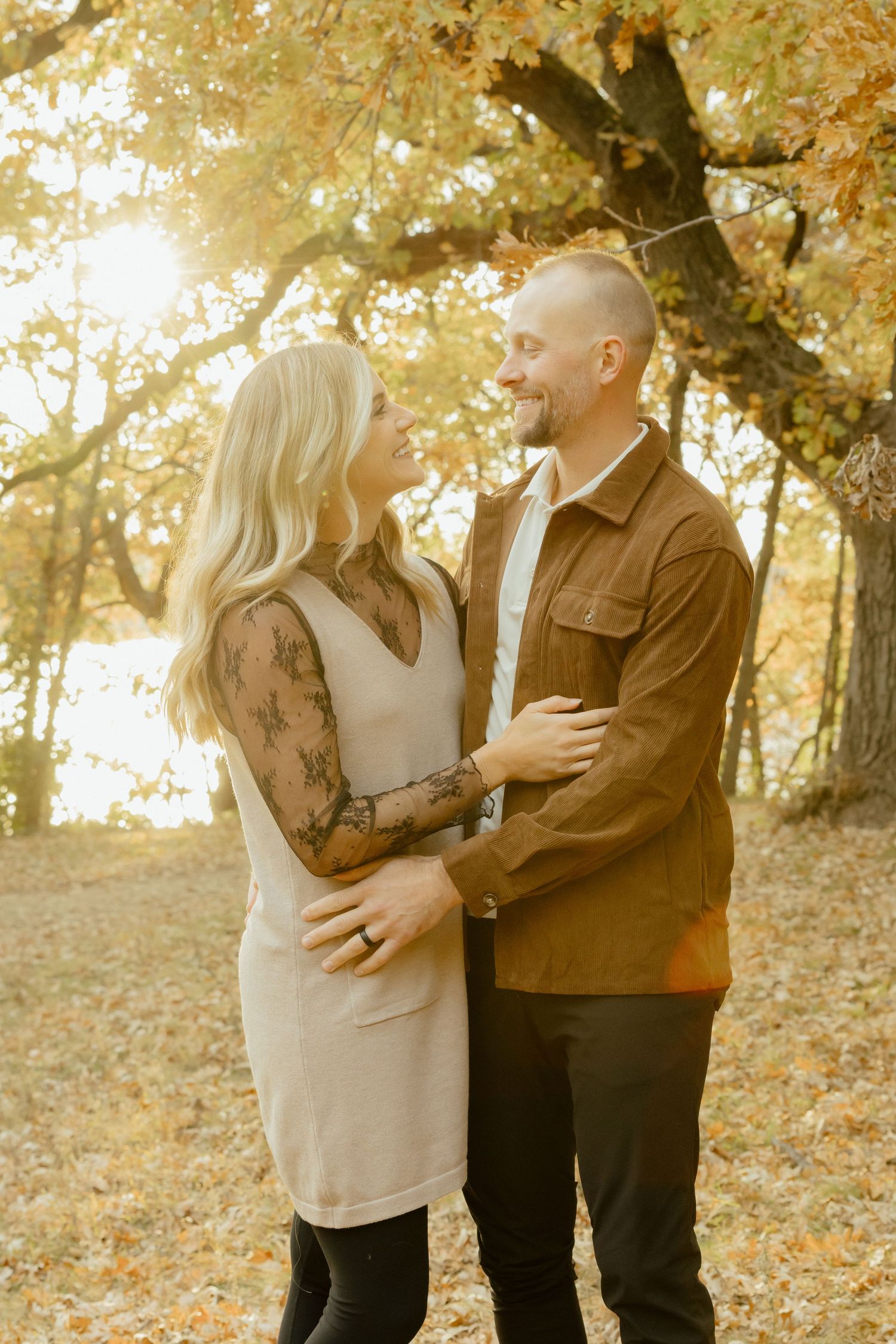 Couple embracing and smiling at each other outdoors among golden autumn trees in warm fall light.