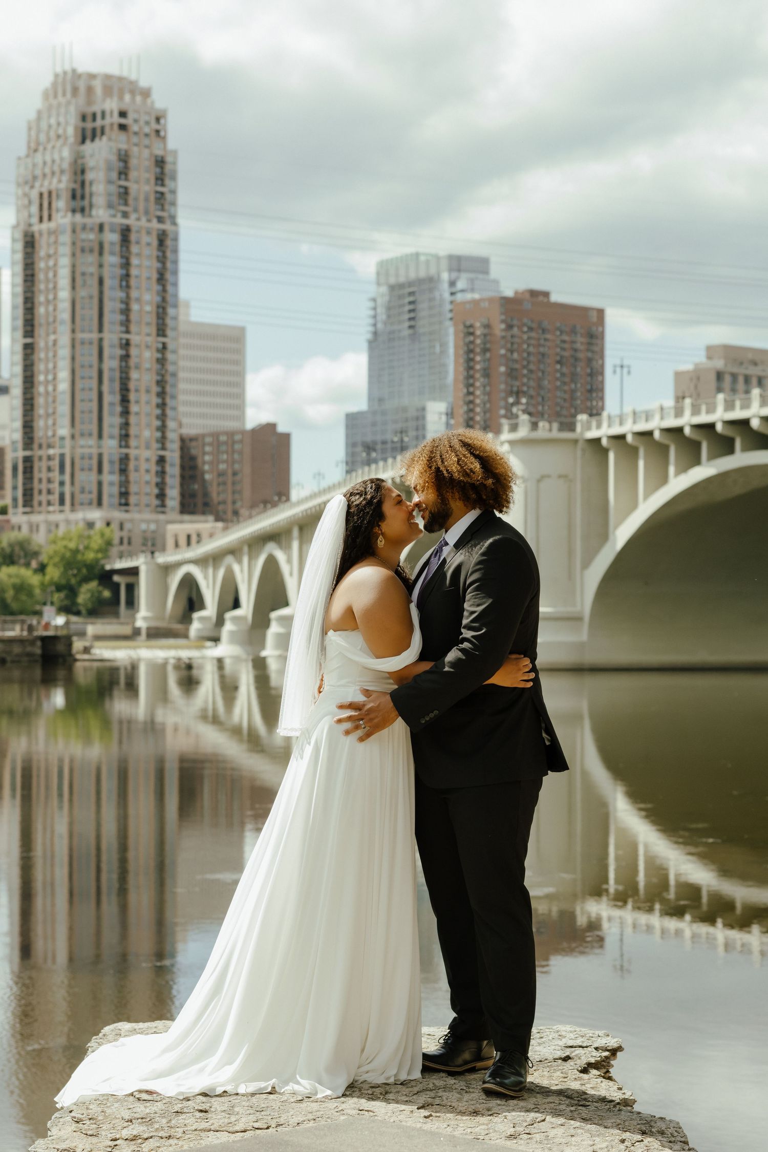 Bride and groom share a romantic kiss by the river with a city bridge and skyline backdrop on their wedding day.