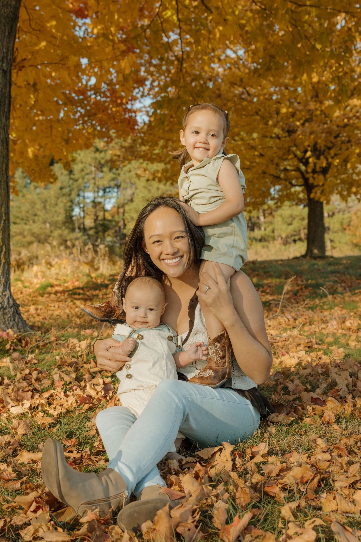 A smiling mother sits in autumn leaves holding a baby while a toddler perches on her shoulders outdoors.