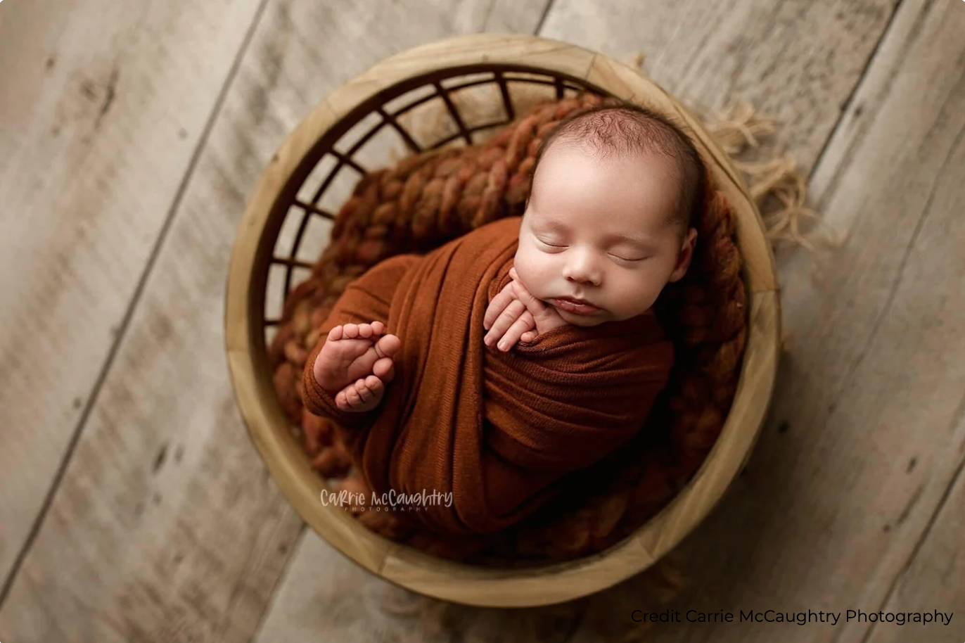 Newborn baby wrapped in rust-orange swaddle sleeping peacefully in a wooden basket with chunky knit blanket.