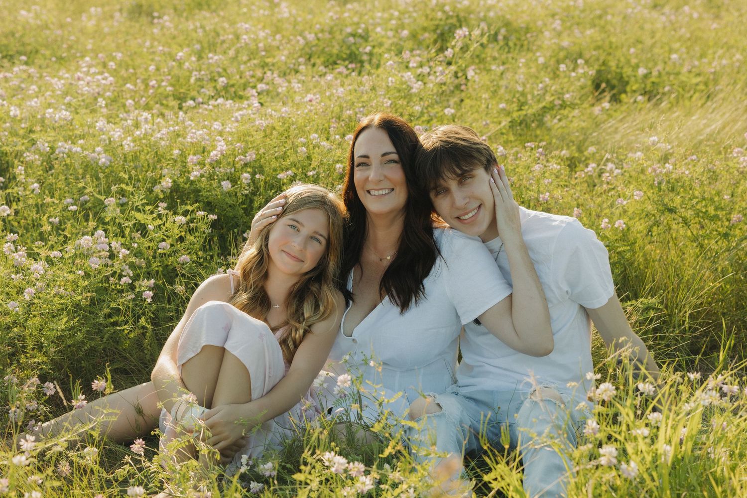 Three smiling women sitting together in a sunny wildflower meadow, wearing white outfits in a bright outdoor portrait.