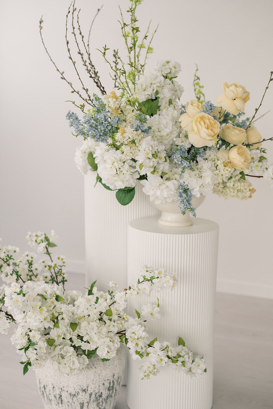 White vases with elegant floral arrangements featuring cream roses, blue and white blooms, and cherry blossoms on white pedestals.