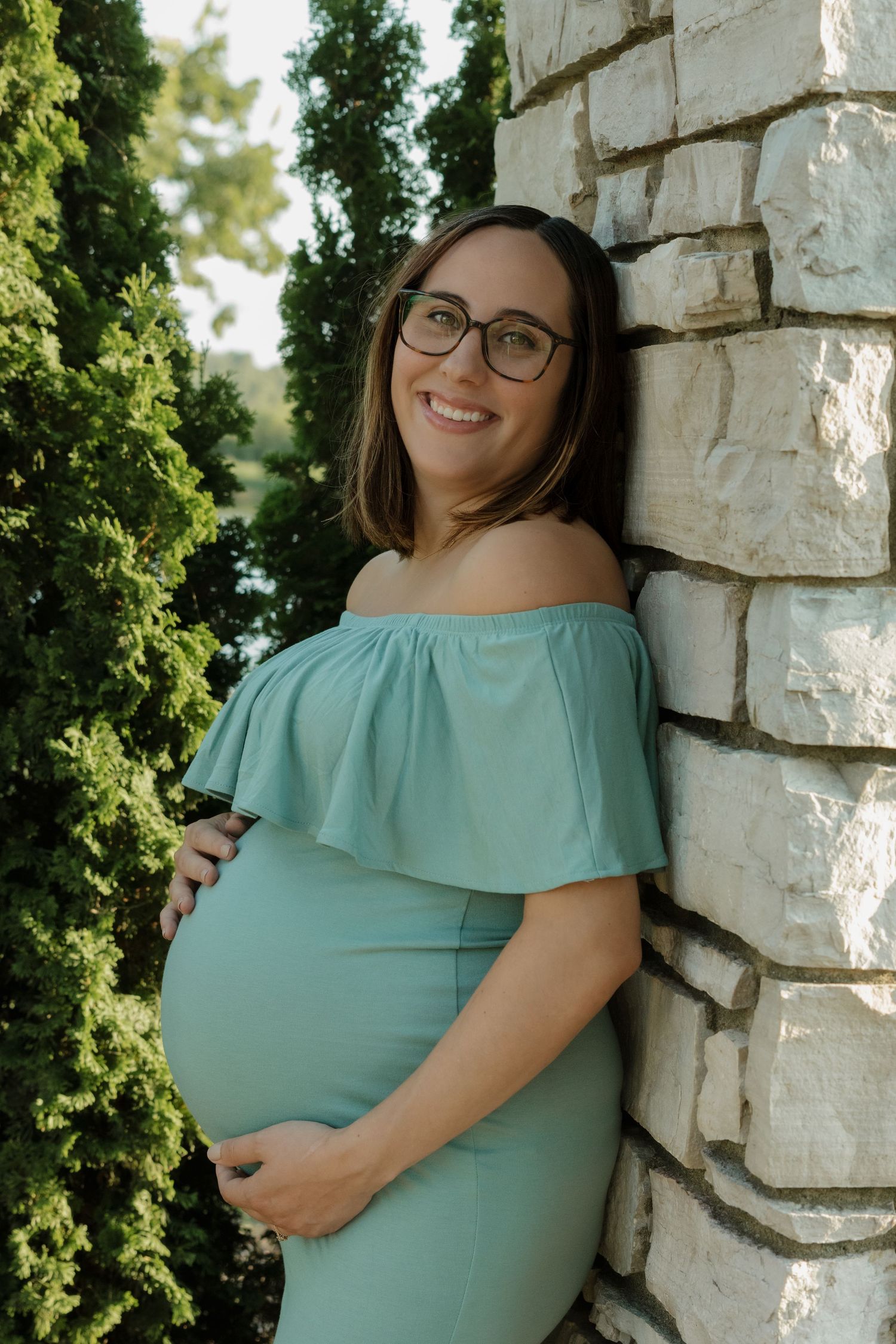 Smiling pregnant woman in teal off-shoulder dress, glasses, leaning against stone wall outdoors.
