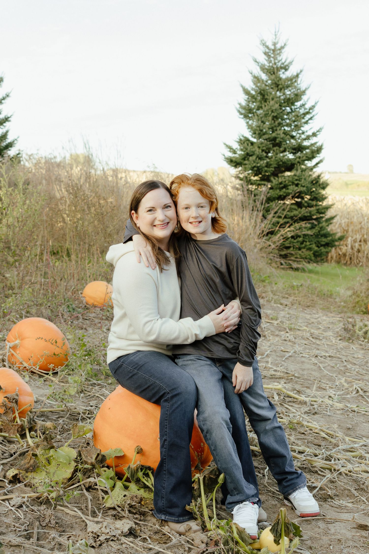 A woman and red-haired child smile together sitting on pumpkins in a fall pumpkin patch with evergreen trees.