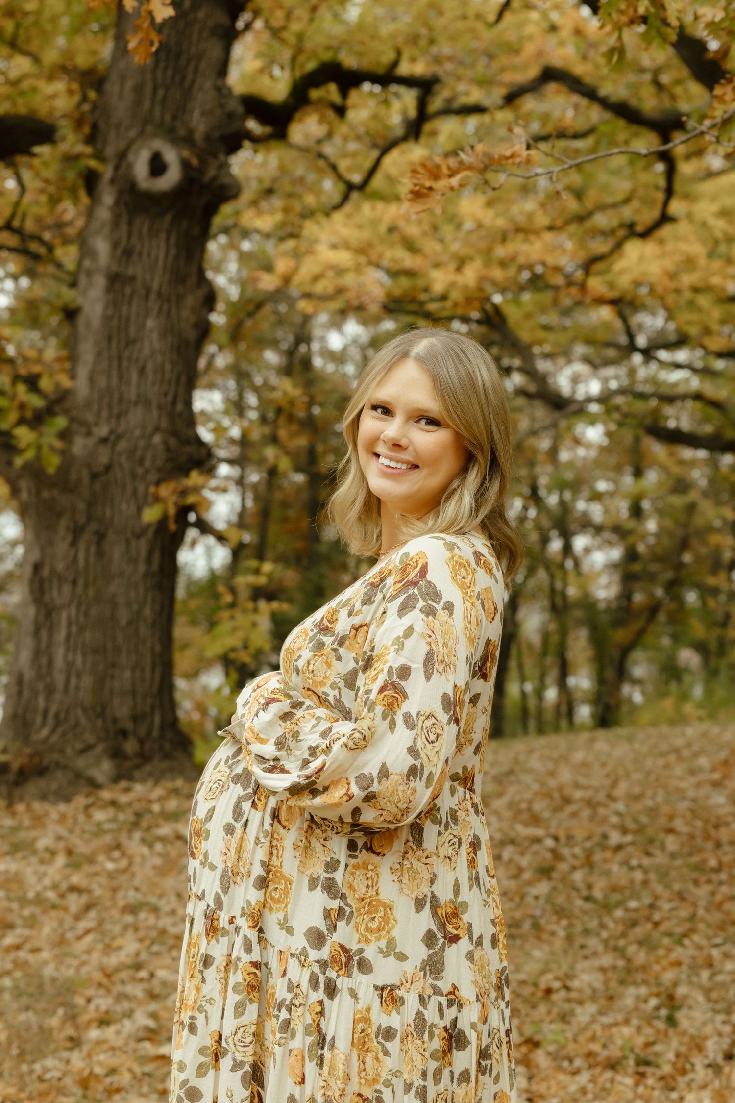 Smiling pregnant woman in floral dress posing outdoors among autumn trees with golden leaves.