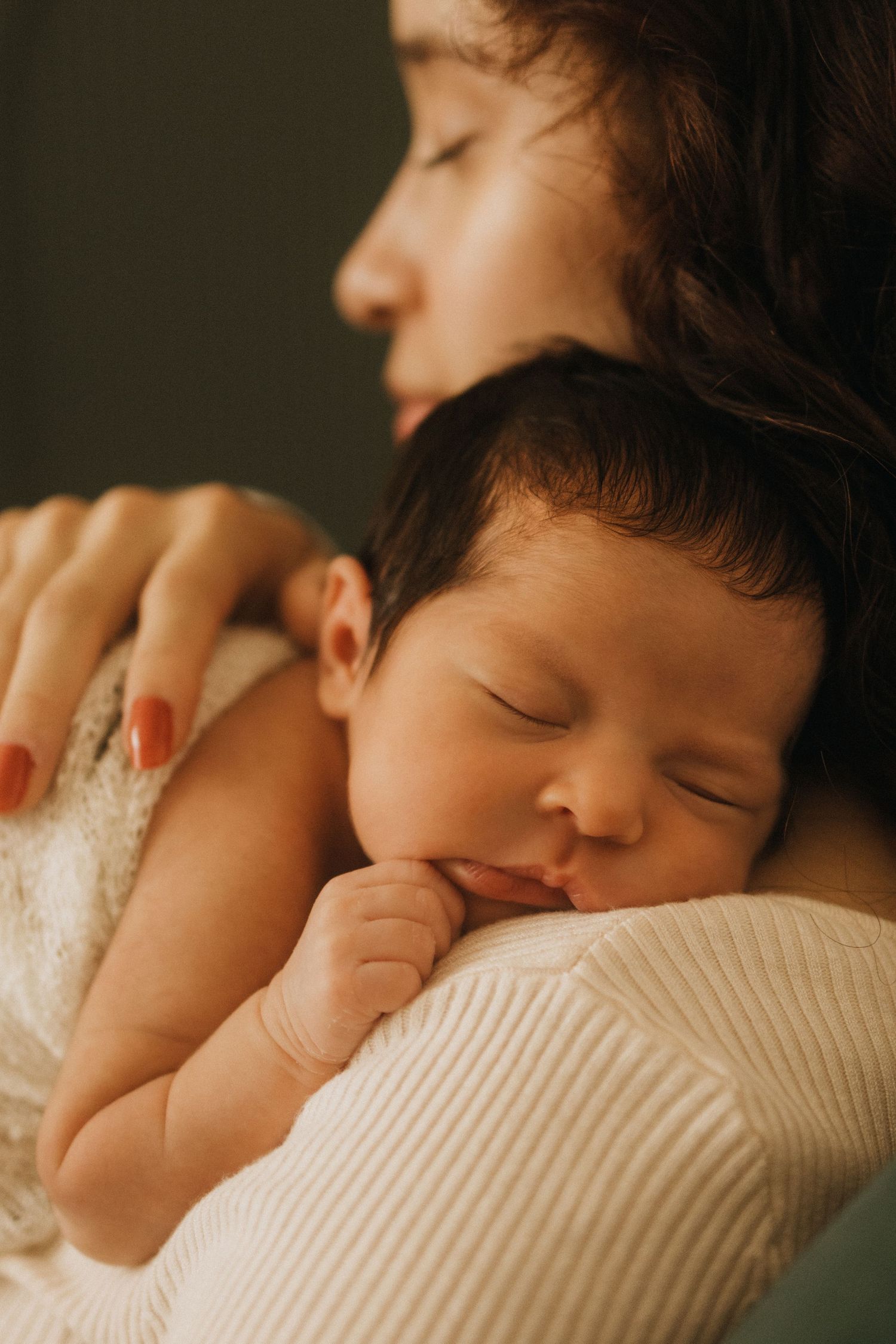 A mother tenderly holds her sleeping newborn baby against her chest in a warm, intimate portrait.