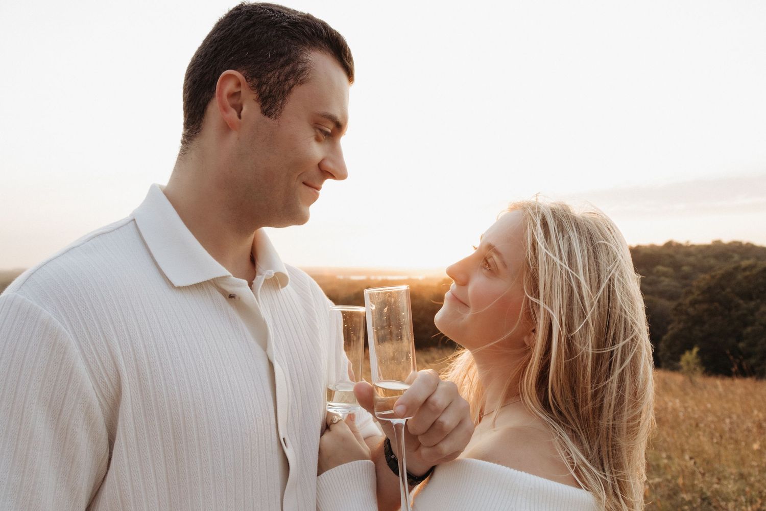 Couple sharing champagne flutes at sunset in a golden field, gazing lovingly at each other.
