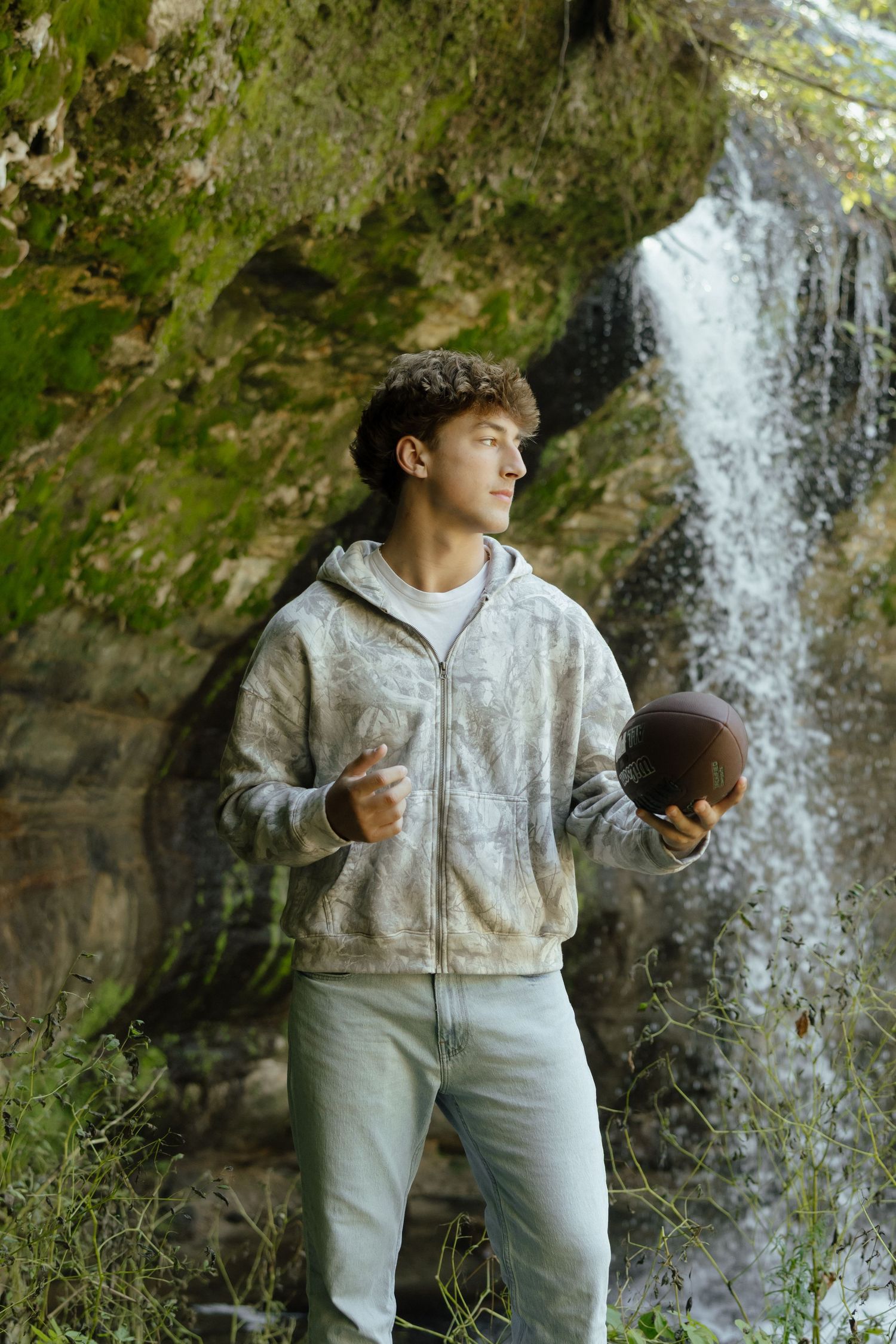 A person in a gray hoodie and light pants poses in front of a cascading waterfall in nature.