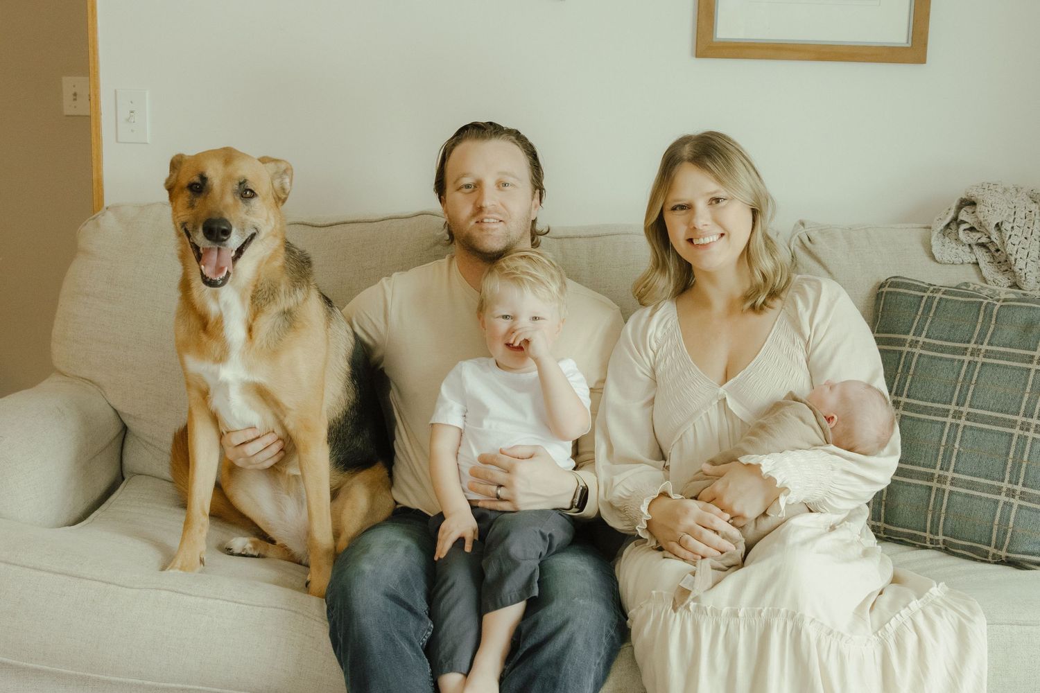 Happy family portrait on couch with large brown dog, man, toddler, woman holding newborn baby in bright living room.