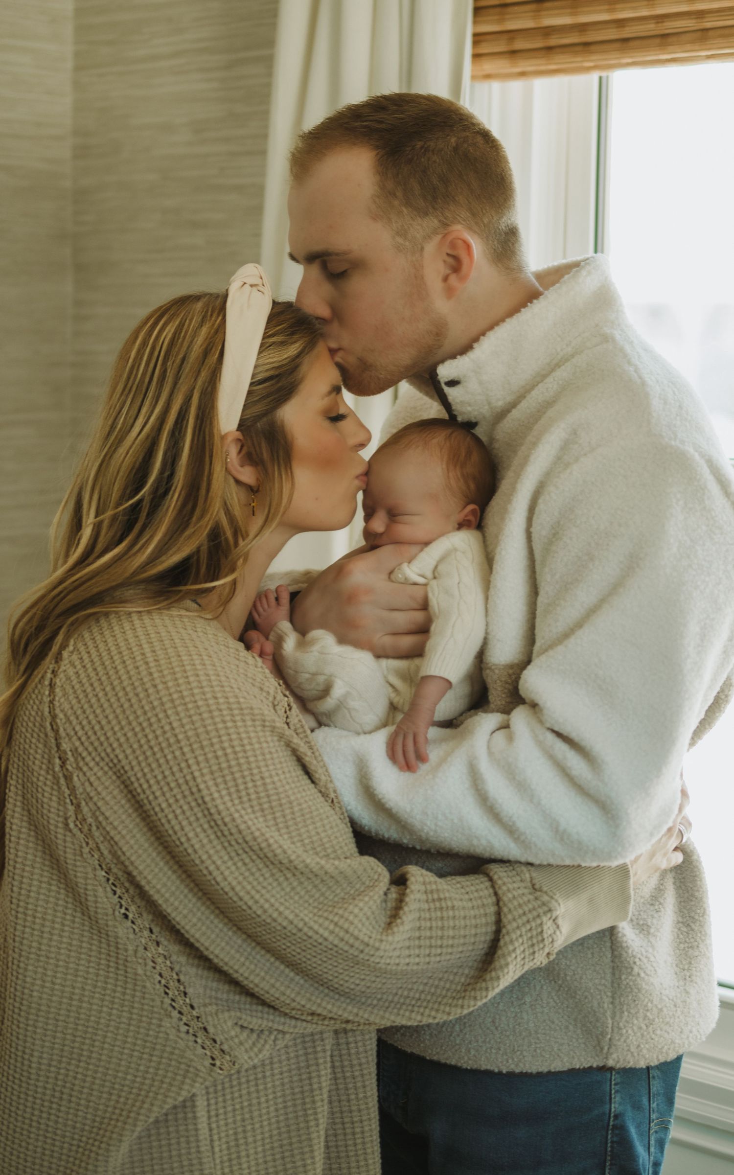 Intimate family moment with newborn baby in soft winter sweaters by window.